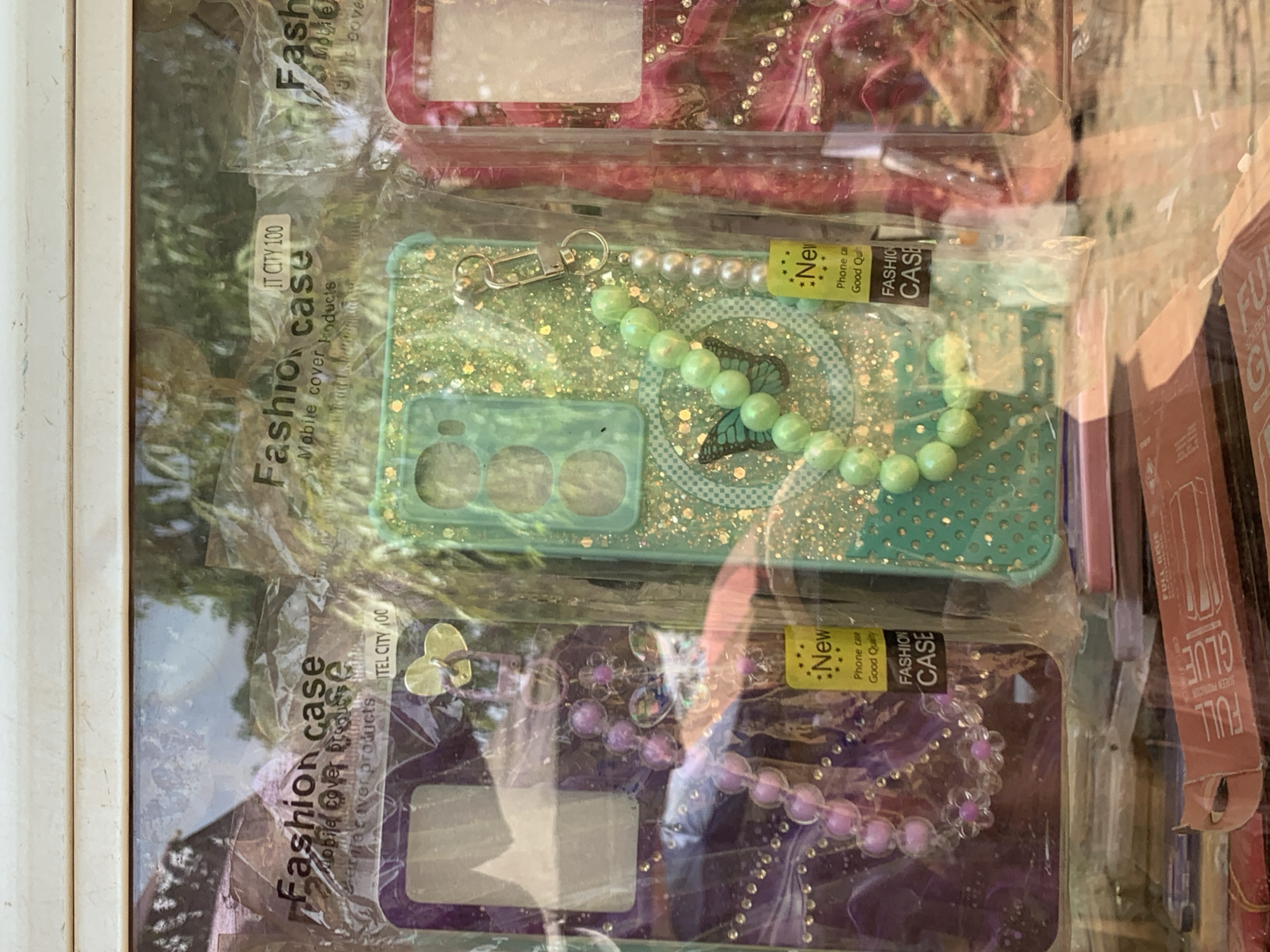 This close-up image presents a display of colorful smartphone cases, likely for sale in a market stall or shop in Ilorin, Nigeria. The cases are individually sealed in clear plastic packaging.
Three main phone cases are visible. The top case is a reddish-pink, embellished with small, glittery accents or rhinestones. Below it, a light green or mint-colored case features a sparkling glitter finish and a central butterfly design. This green case is accessorized with a string of matching light green beads and some pearl-like beads, attached with a metal clasp. The bottom case is purple, also with a glittery texture, and includes a purple beaded strap and a small golden heart-shaped charm.
Visible text on the packaging provides product information. Labels repeatedly feature "Fashion case," "Mobile cover products," and "IT CITY 100." Yellow tags on the middle and bottom cases state "New," "Phone case," and "Good Quality," along with "FASHION CASE." To the right, partial packaging for "FULL GLUE SCREEN PROTECTION" indicates other phone accessories are available. The scene is brightly lit, suggesting daytime.