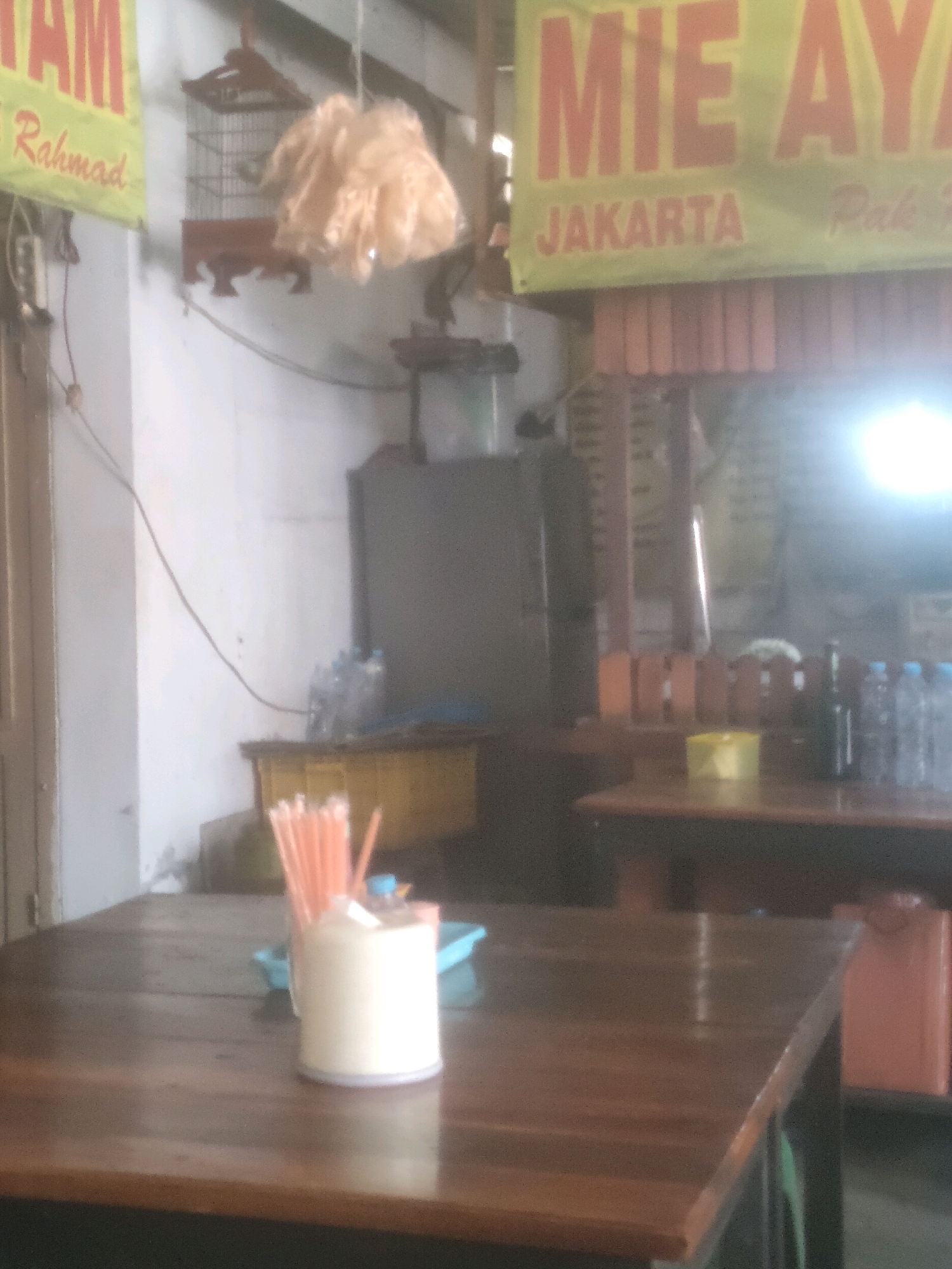 The image depicts an interior scene that appears to be a casual eatery or food stall, possibly located in Surabaya, Indonesia, suggested by the text on the banners. In the foreground, a wooden table is visible, with a white ceramic container holding orange straws and a blue tray with some condiment containers on it. The table surface is wooden and shows signs of wear.
In the background, there are several elements that contribute to the atmosphere of the establishment. A large, green banner with red lettering that reads "MIE AYAM" (Chicken Noodles) and "JAKARTA" is prominently displayed. Below this, another banner with yellow lettering is partially visible. A birdcage hangs from the ceiling, with a cluster of what looks like fried food or snacks in a plastic bag suspended above it. To the right, a stainless steel refrigerator is positioned against a white wall. Further back, more wooden furniture, possibly tables and chairs, and shelving units can be seen. Bottles of water are lined up on a shelf or counter on the right side of the image. The lighting in the scene is somewhat dim and uneven, with a bright light source, possibly a fluorescent lamp, creating a glare on the right side of the image. The overall impression is of a functional, no-frills dining space.