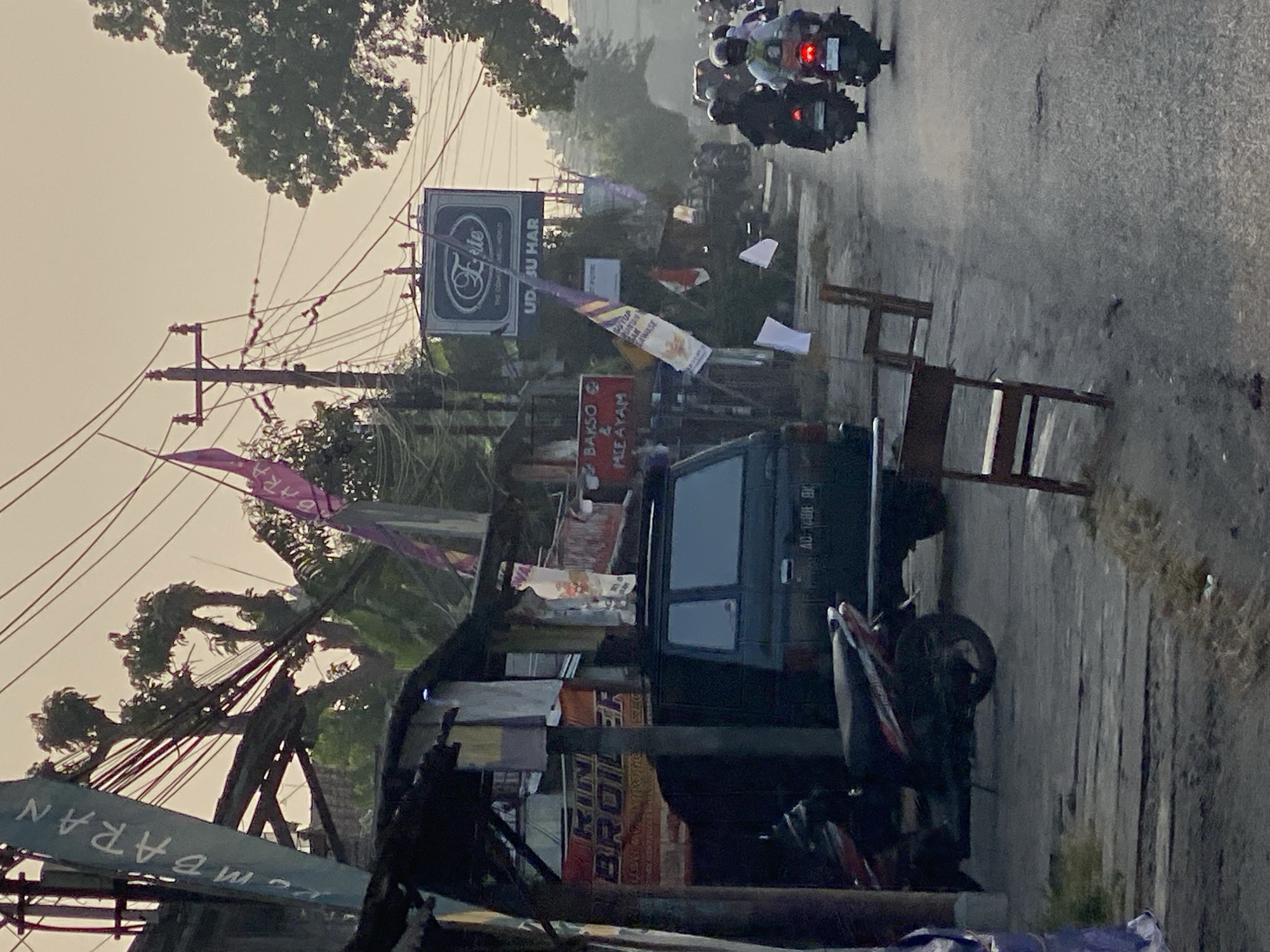 The image depicts a street scene in Kasihan, Indonesia, likely during the early morning or late afternoon, given the soft, diffused lighting. The foreground features a dark-colored motorbike parked on a wooden plank walkway, beside a dark-colored utility vehicle. To the left, a blue and white banner with Indonesian text is partially visible. Further back, a small storefront with a red sign displaying "RINK BROILER" is evident. Above the storefront, a collection of utility poles and wires dominate the upper portion of the frame, with various flags and banners attached. One sign reads "Elite THE COMFORT WORLD" with smaller text below that is unreadable. Another red sign shows "BAKSO & MIE AYAM" (meatball and noodle soup). The street itself is paved and shows signs of wear. Several wooden chairs are placed along the side of the road. In the background, a procession of motorbikes, some with passengers, travels down the street. The overall atmosphere is that of a bustling, everyday street in a rural or semi-urban Indonesian setting.