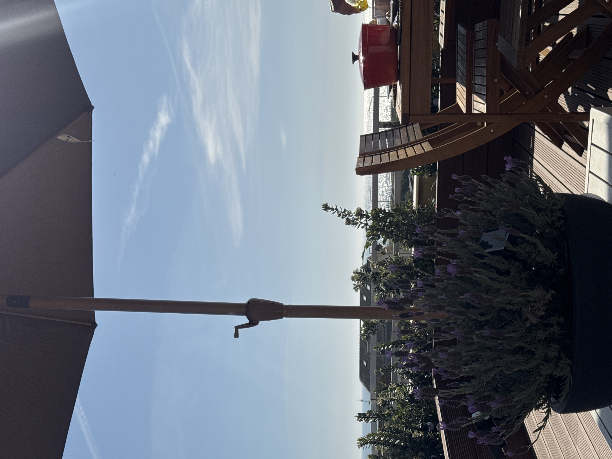 The image depicts an outdoor scene in Vila Nova de Gaia, Portugal. A patio is visible, furnished with wooden furniture including chairs and a table. A large black flower pot containing lavender plants sits on the edge of the patio. A red cooking pot rests on the table. An open brown patio umbrella is partially visible in the left and center foreground. The Atlantic Ocean is visible in the background. The sky is clear blue with some wispy clouds. Residential buildings are visible in the midground.