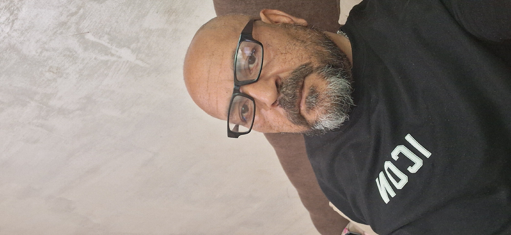 An adult male subject is depicted from the mid-torso upwards, situated in the city of Simeonovgrad, Bulgaria. The individual is bald and wears dark, rectangular-framed eyeglasses. He possesses a full beard and mustache, predominantly grey in color. He is wearing a black crew-neck shirt or sweatshirt, featuring the word "ICON" embroidered in light grey or white capital letters on the upper left chest area. A metallic chain is visible around his neck. The subject is positioned facing forward, with his gaze directed towards the camera. He appears to be seated or leaning against a brown upholstered surface, which is partially visible on the right side of the frame. The background consists of a plain, light-colored wall with a subtle textured finish, exhibiting minor smudges and faint linear marks.