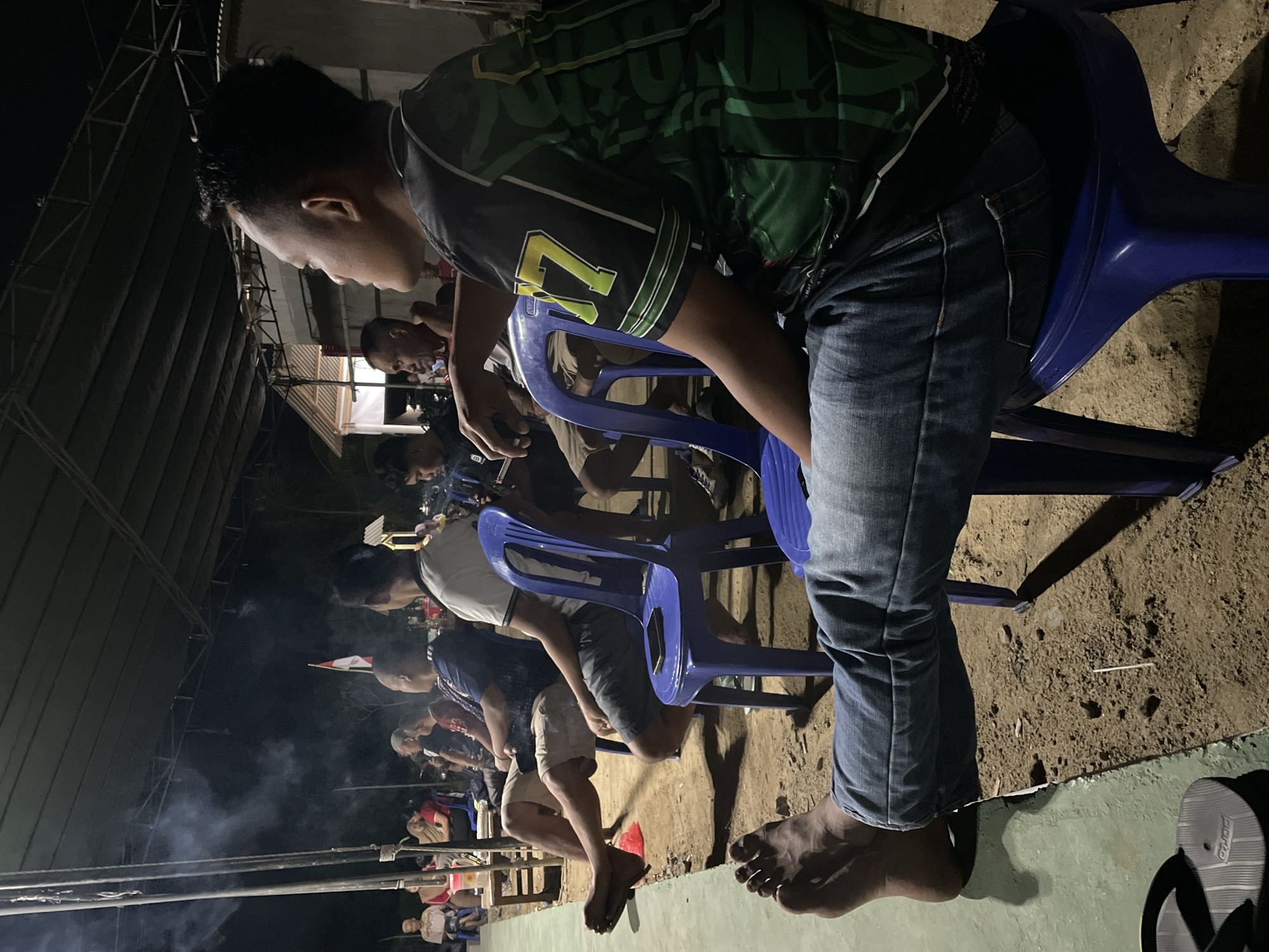 The image captures an outdoor gathering at night, likely in Kagungan Ratu, Indonesia, with an audience seated on chairs under a canopy. In the foreground, a man wearing blue jeans and a black and green patterned shirt with the number "17" visible on the sleeve is seated on a blue plastic chair, his legs extended. To his left, another person's bare feet and legs in blue jeans are visible, resting on a green surface.
In the background, several other individuals are seated, some facing forward, and others turned towards something out of frame. There is a small Indonesian flag displayed. Smoke is visible rising from the left side of the frame. The ground appears to be a mix of dirt and sand. The scene suggests a community event, possibly a performance or gathering, taking place outdoors at night.