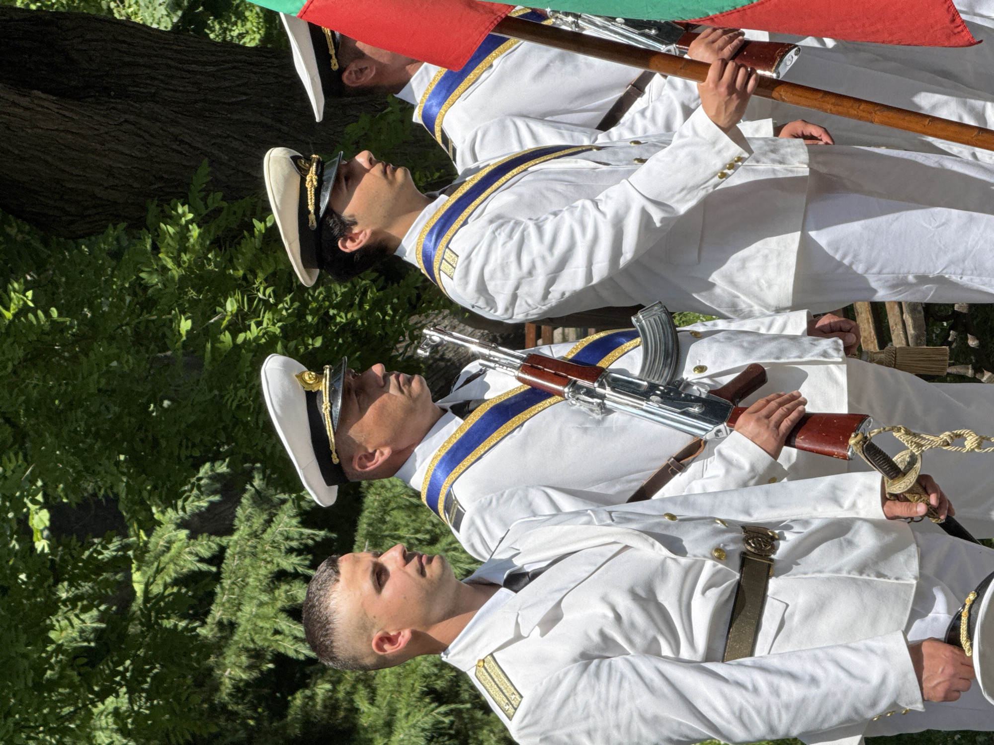 A group of at least three men, dressed in formal white military or naval-style uniforms, are captured in an outdoor, brightly lit setting. The image is rotated, showing the men standing upright in a line, looking towards the right side of the frame.
The men wear white jackets with gold buttons and detailing. The man in the foreground has gold shoulder boards, while the two men behind him wear blue and gold sashes across their chests. Two of them wear white peaked caps adorned with gold trim and insignia.
Each man carries ceremonial items: the man second from the front holds what appears to be a Kalashnikov-style rifle with a wooden stock and metallic magazine. The third man also holds a long rifle with a wooden stock. The man in the foreground carries a decorative sabre or sword with a gold hilt and a rope tassel, and wears a brown belt with a prominent buckle. Partially visible at the top of the frame is a flag with red, green, and white stripes, consistent with the national flag of Bulgaria, held by one of the men.
The background consists of lush green foliage and tree trunks, indicating a daytime outdoor event. The bright sunlight suggests clear weather. Given the uniforms, ceremonial weapons, and national flag, the men are likely participating in a parade, official ceremony, or guard duty in Burgas, Bulgaria. Their expressions are formal and stoic, fitting for such an occasion. No visible text is present in the image.
