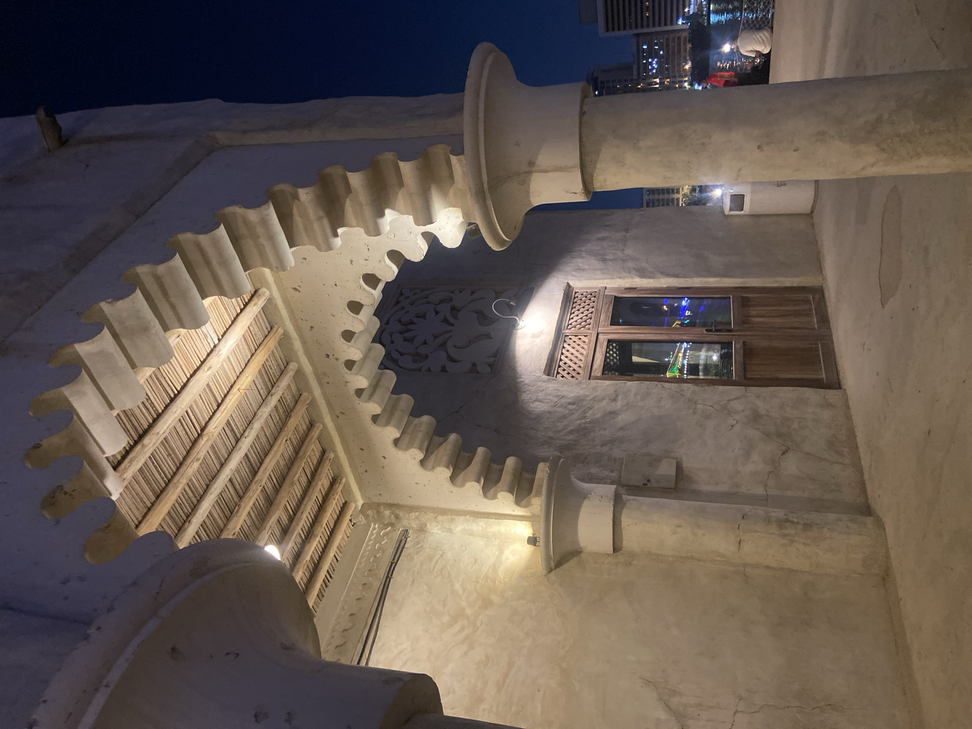 The image depicts an outdoor view at night in Deira, United Arab Emirates. The foreground features a building entrance with a decorative, scalloped archway, rustic wooden double doors with glass panels, and a rough, textured wall. The ceiling above the entrance is constructed with wooden beams and palm fronds. A large, cylindrical column is visible to the right of the entrance, and further to the right, a white trash receptacle stands near the edge of a paved area. In the background, a body of water reflects the lights of buildings and a few people are seated by the water's edge. The overall scene suggests a historical or culturally themed area within the bustling city.