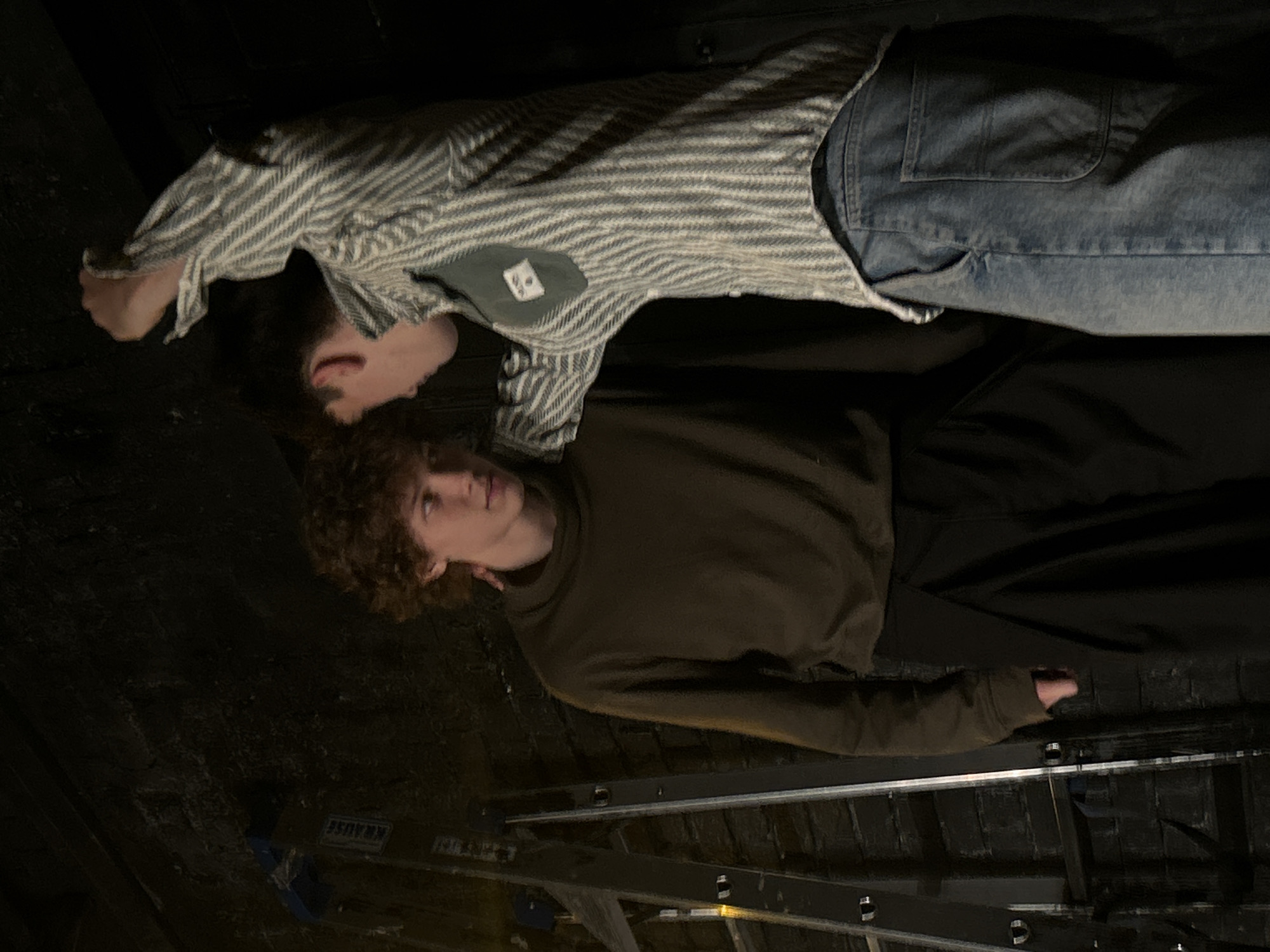 Two young men are standing in front of a dark brick wall with a ladder to the left of the frame. The man on the left has curly brown hair and is wearing a dark brown turtleneck and black pants. The man on the right has short dark hair and is wearing a striped shirt and blue jeans. He has his arm around the man on the left's shoulder and is looking down at him. The scene appears to be indoors, possibly in a studio or backstage area, given the presence of the ladder and the dark, raw aesthetic of the background.