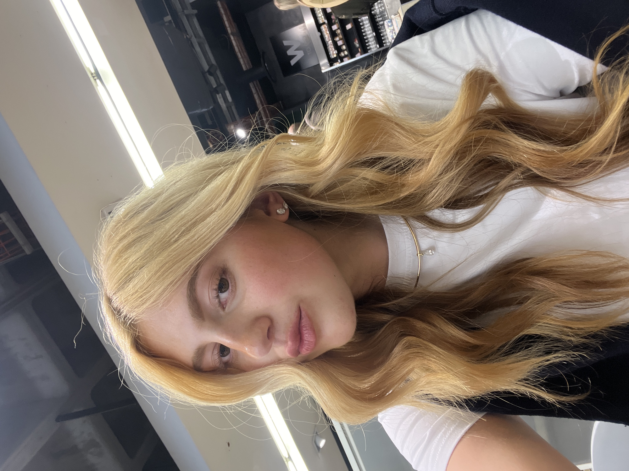 A medium shot of a blonde-haired woman is captured indoors. Her long, wavy hair cascades over her left shoulder. She is wearing a white t-shirt. An earring is visible in her left ear. A thin gold necklace adorns her neck. The background reveals an interior, possibly a store or salon. A display with various items is noticeable on the left. Given the presence of display shelves and the items shown, it's possible this image was taken inside a shop in Johannesburg.