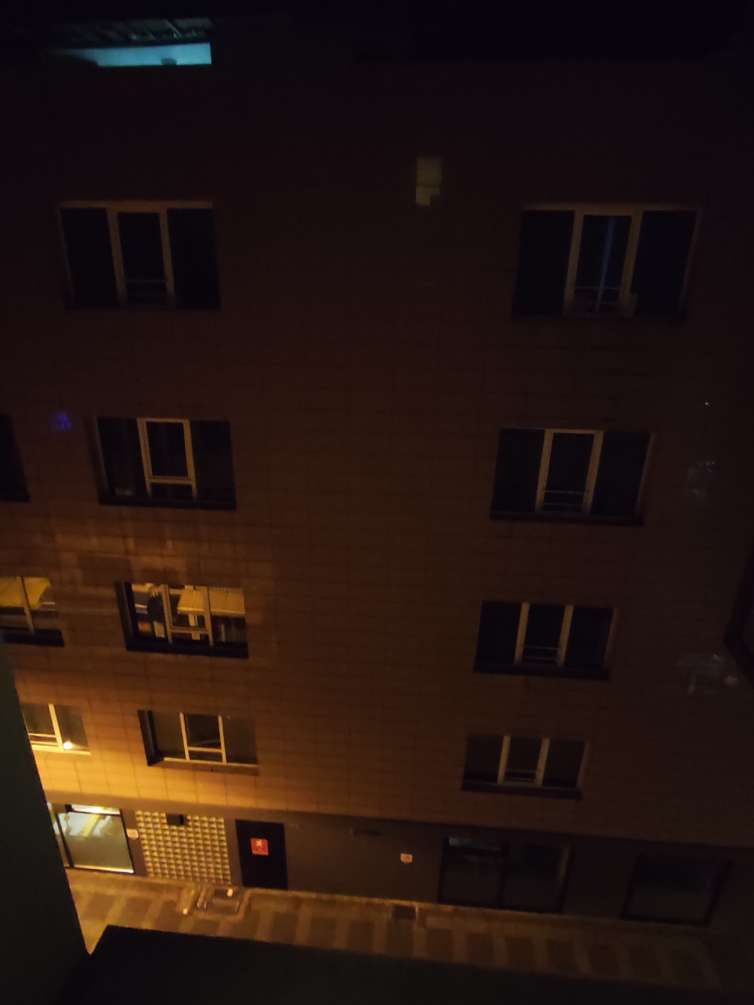 This image depicts a building at night, likely in Kadıköy, Turkey. The perspective is from a higher vantage point looking downwards at multiple levels of windows. The windows appear to be multi-paned, with some illuminated from within and others dark. The building's facade is a textured brick or stone. In the lower section of the image, there is an entrance area with doors and what appears to be a patterned wall. The overall lighting suggests it is nighttime or very dimly lit. There are no people or discernible actions visible.