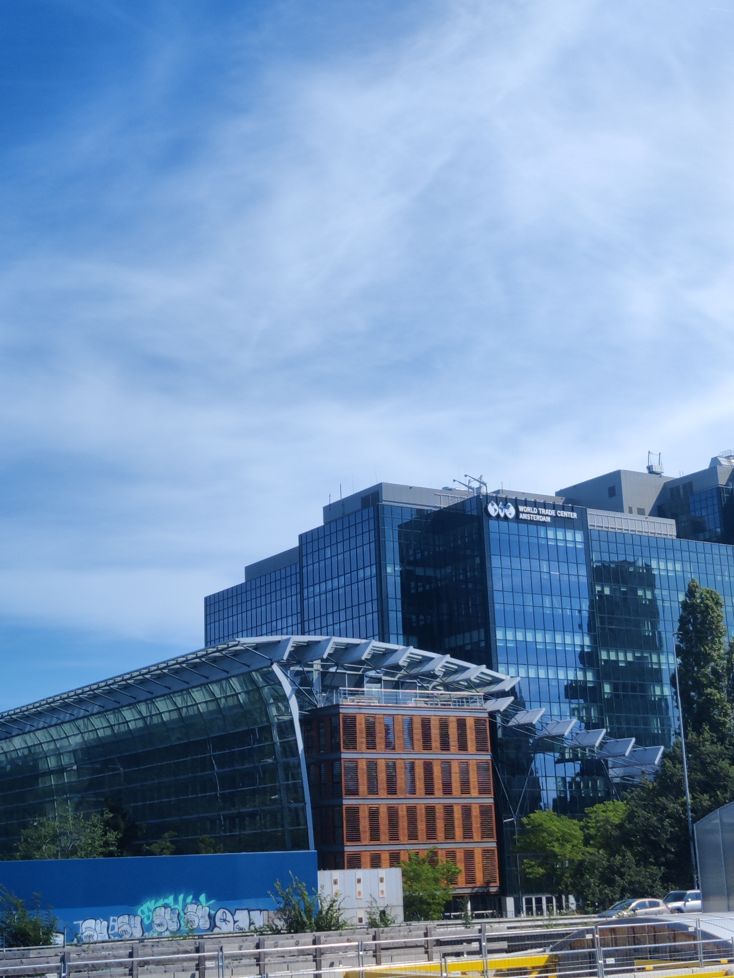 The image shows a modern glass-fronted building in Amsterdam, Netherlands, with "WORLD TRADE CENTER AMSTERDAM" visible on the facade. The building is blue and glass, with a curved glass structure in the foreground. A smaller, brown building with wooden shutters is next to the curved structure. Trees and greenery are visible around the buildings. The sky is blue with some white clouds. In the foreground, there is a blue wall with graffiti and a metal fence. A road with a couple of cars is also visible in the background.