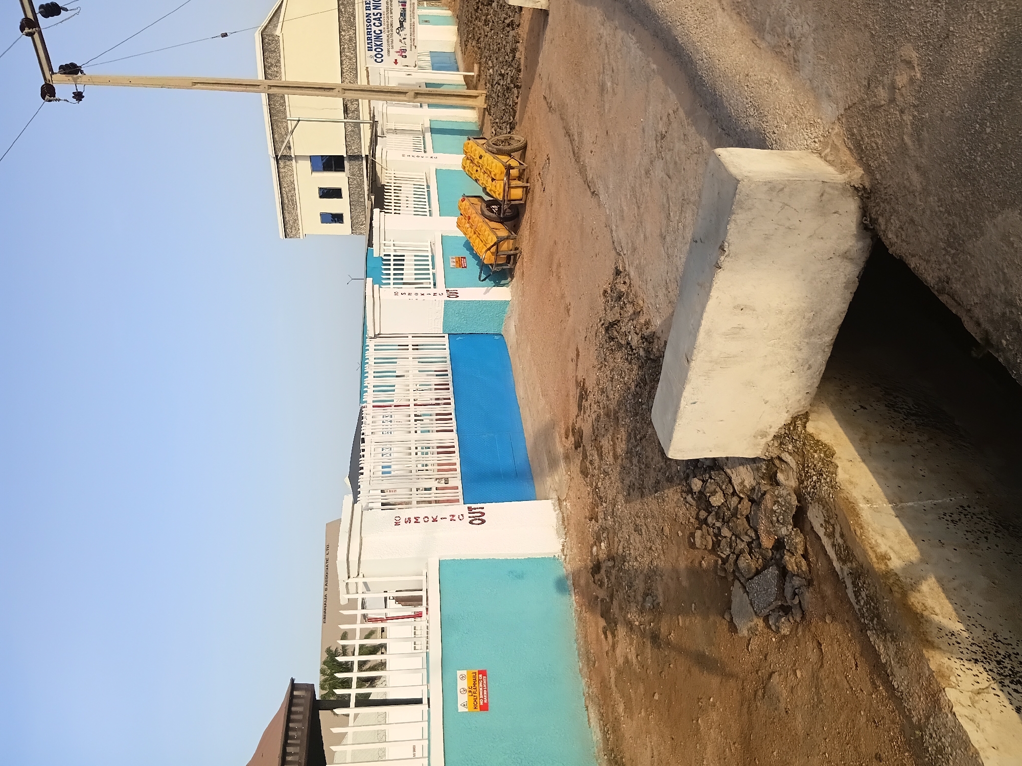 The image shows a street scene in Jalingo, Nigeria, with a clear blue sky overhead. The foreground features a partially constructed concrete structure, possibly a bridge or culvert, with exposed rebar and rough terrain surrounding it. To the right, a utility pole with electrical equipment stands tall.
In the midground, a bright blue and white building with modern architecture is visible, featuring multiple windows. Adjacent to it is a fenced area with white picket fences and a blue wall. Several yellow jerrycans filled with liquid are placed on carts just outside this fenced area. A sign on the blue wall reads "NO SMOKING OUT" in red lettering.
Further to the left, another building with a brown roof is partially visible, also behind a white fence. A large sign on the blue wall bears a warning symbol and text indicating "HIGHLY FLAMMABLE" and "NO SMOKING OR OPEN LIGHTS". The ground in this area is predominantly sandy and dusty, with some scattered rocks and debris. The overall impression is an urban environment, possibly an industrial or commercial zone, with some ongoing construction or maintenance work evident.