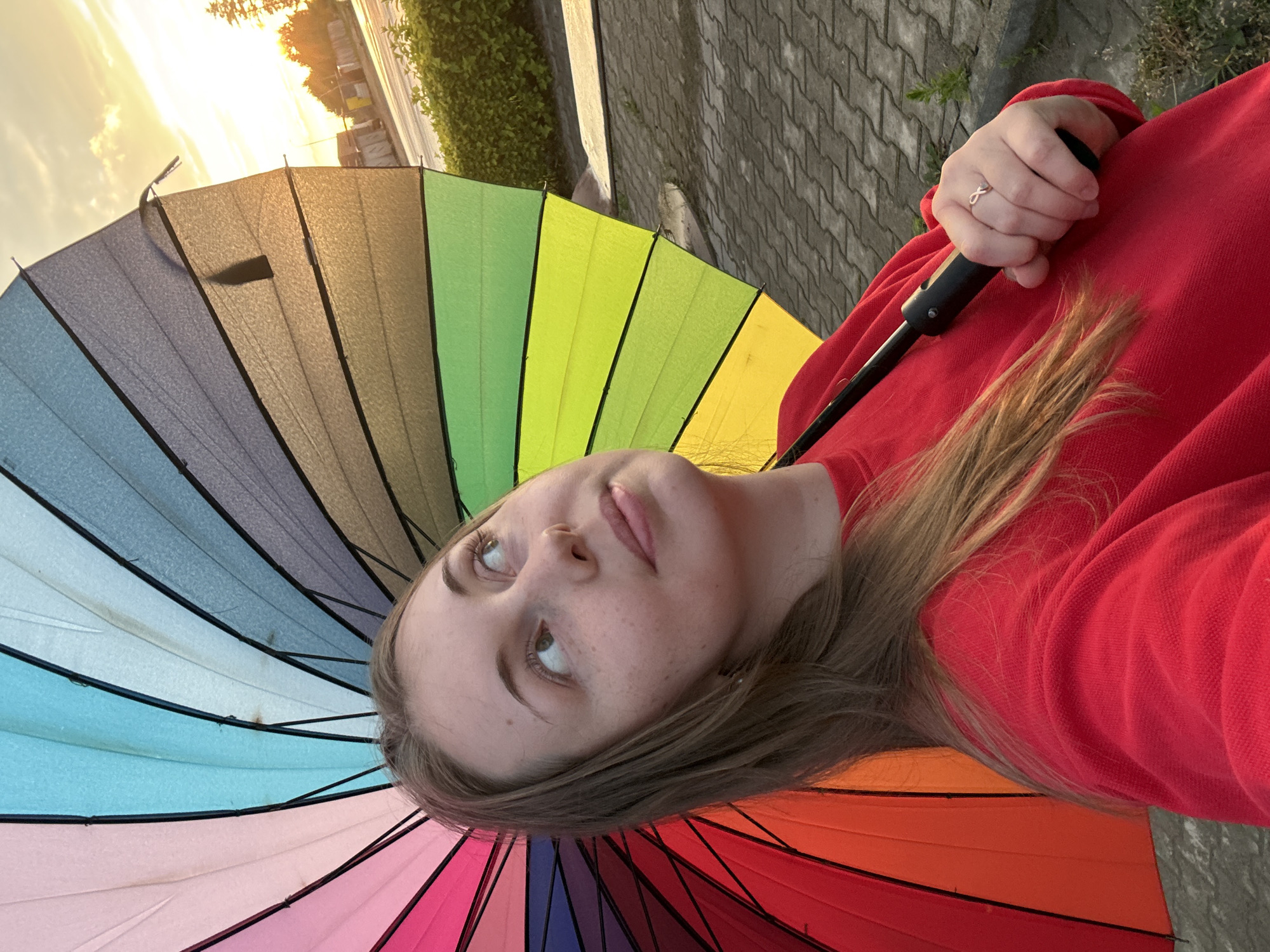 In an outdoor, paved urban setting under a sunset sky, a young woman with light brown hair and freckles holds a large, open, rainbow-colored umbrella. She wears a red crewneck sweatshirt. The umbrella's spokes are black, and the fabric panels transition through shades of pink, purple, blue, green, yellow, and orange. The woman's right hand grips the black handle of the umbrella, and a silver ring with an infinity symbol is visible on her ring finger. The background shows a paved ground, green bushes, and a street with buildings and trees illuminated by the setting sun.