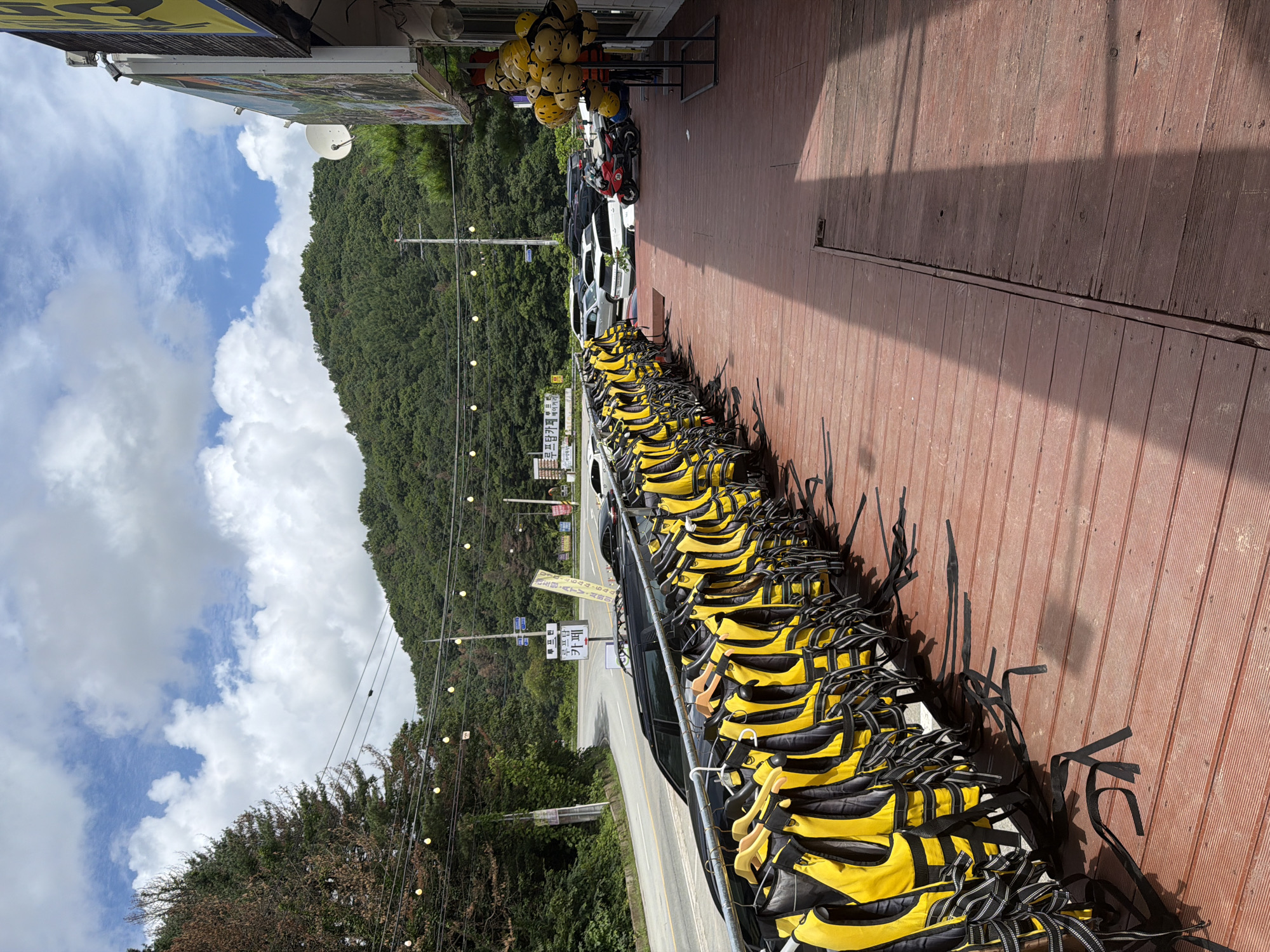 The image shows an outdoor scene near a road in Seo-myeon, South Korea, with a focus on a row of bright yellow life jackets hanging on a metal rack in the foreground. The life jackets are neatly arranged, each on a wooden hanger, suggesting they are available for rent or use. Behind the life jackets, a wooden deck extends into the frame, with shadows indicating the presence of overhead structures.
A road curves through a lush green hillside in the background, with cars parked along the roadside and on a lot to the right. A building with a prominent sign, partially obscured, is visible on the right side of the image. There are strings of lights strung across the scene, and a stack of yellow helmets is perched on a stand near the building. The sky above is partly cloudy with patches of blue. No people are visible in the image. The overall impression is that of an establishment preparing for or situated near outdoor recreational activities, possibly water sports, given the presence of the life jackets and helmets.