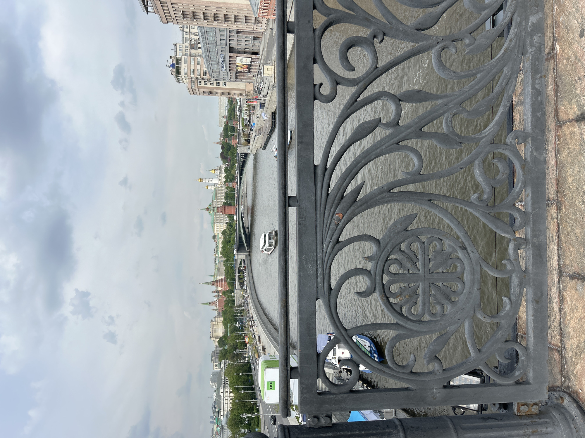 The image depicts a view of a river in an urban setting from a bridge. The bridge has an ornate metal railing in the foreground with decorative floral and circular patterns. In the background, there's a river with boats and buildings along its banks. Some of the buildings have architectural features reminiscent of the Kremlin in Moscow. The sky is overcast with grey clouds. The location appears to be Moscow, Russia.