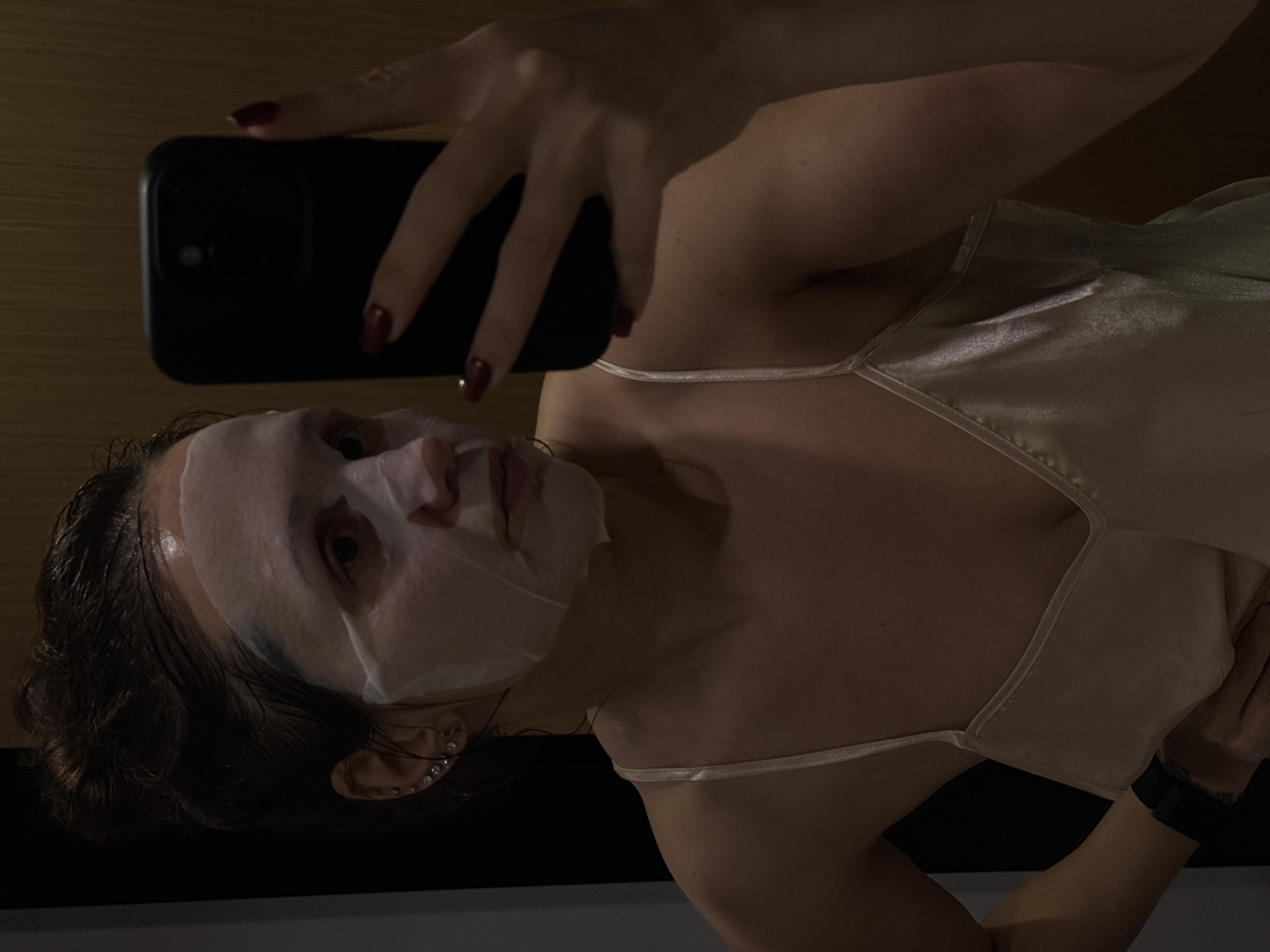 A woman wearing a facial mask is taking a selfie with a black smartphone. She is wearing a light-colored, satin tank top. The background is dark, with a hint of wood paneling visible. A black watch is on her left wrist. The location is not discernible from this image and there is no indication this photograph was taken in Dubai or anywhere else.