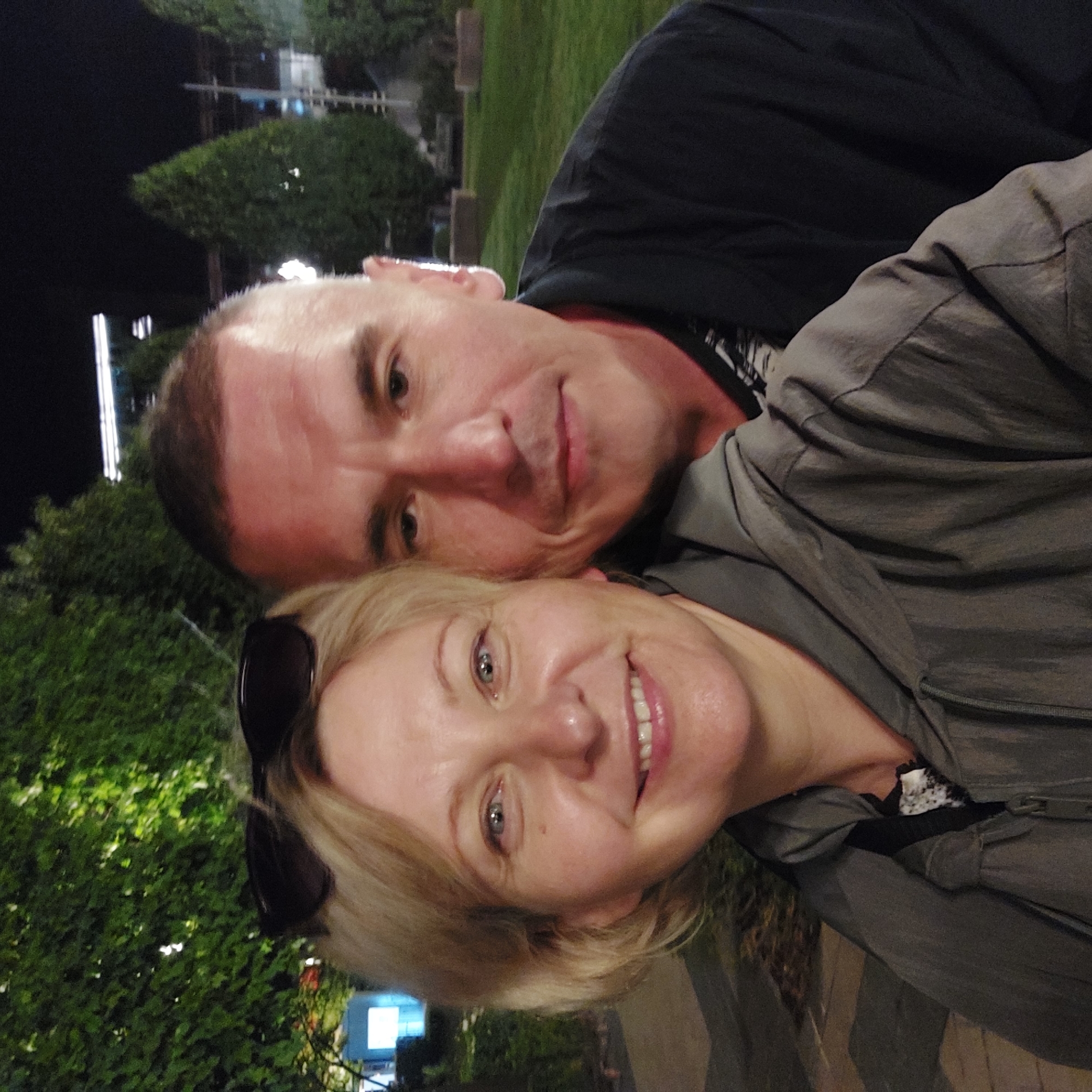 A close-up, front-facing selfie shows a man and a woman standing outdoors at night. The woman, on the left, has blonde hair and is wearing sunglasses on top of her head and a grey hooded jacket. She is smiling broadly. The man, on the right, has short brown hair and is wearing a dark jacket. They are both looking towards the camera. The background shows green trees and some lights, suggesting a park or outdoor public space. This photograph was taken at the Palas Mall park in Iași, Romania.