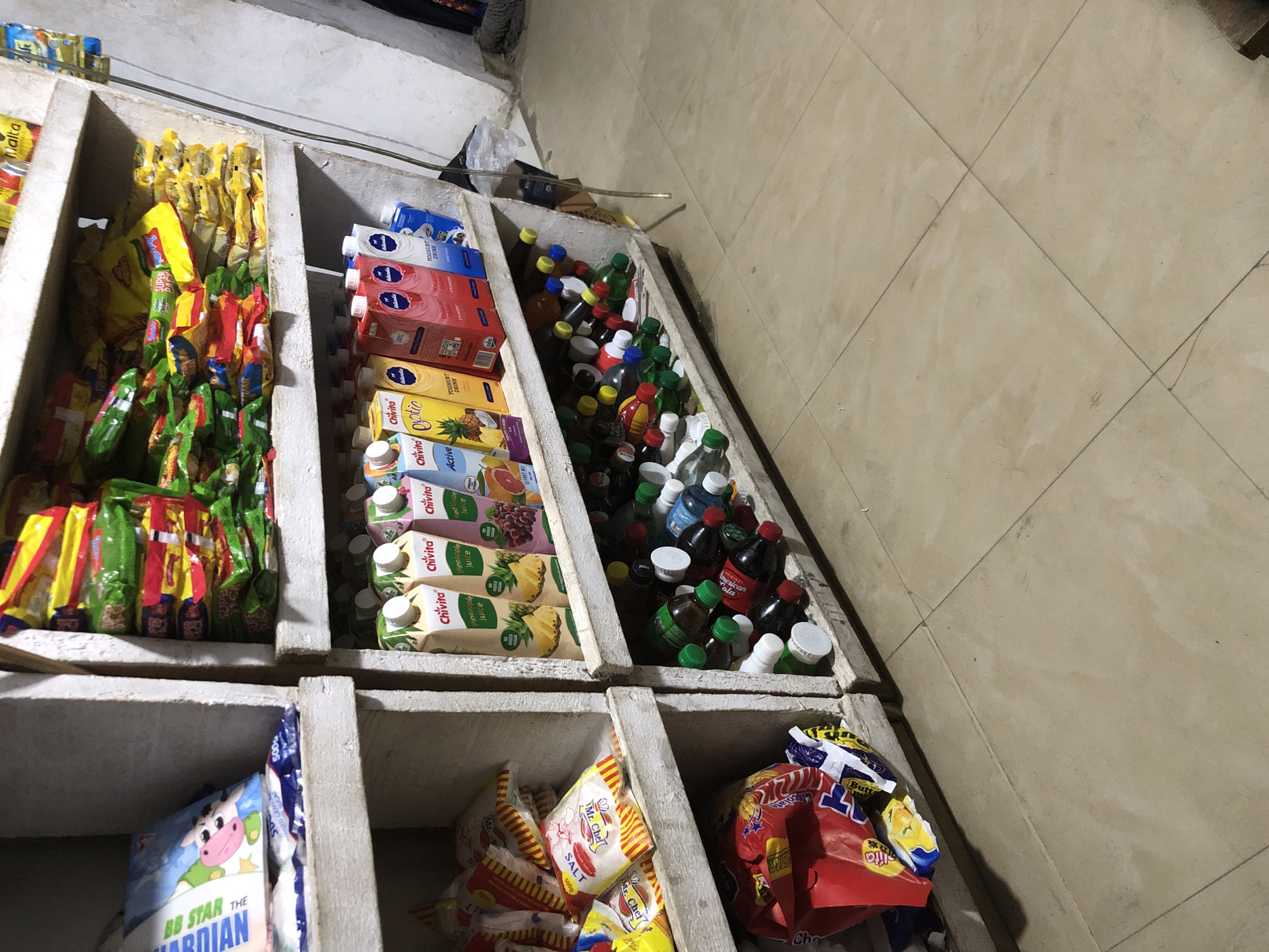 The image shows a shelf in a small shop, possibly a convenience store or a local market stall, in Ado, Nigeria. The shelves are made of wood and are filled with various packaged food items and drinks. On the top shelf, there are stacks of brightly colored packaged snacks and what appear to be instant noodles. On the middle shelf, there are cartons of Chivita juice and yogurt drinks, with labels indicating flavors like pineapple, exotic, strawberry, and red grape. The bottom shelf is crowded with various bottles of soft drinks, some with green caps and others with white, red, and blue caps. A bag of Mr. Chef salt is visible on the left side of the middle shelf. The overall environment appears to be dimly lit and somewhat cluttered, typical of a small retail space in Nigeria. The floor is tiled with light-colored square tiles. There are no people visible in the image.