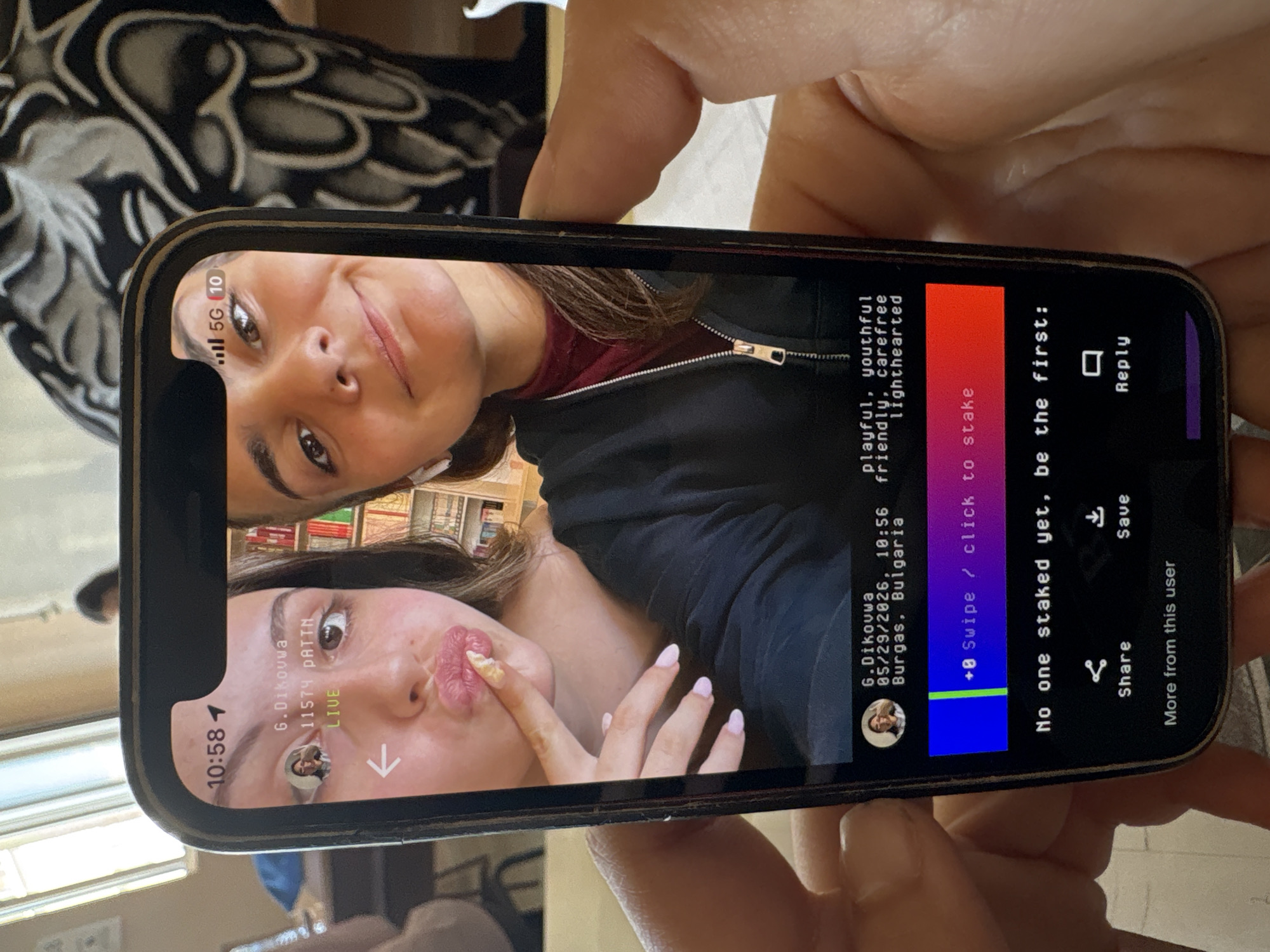 The image shows a person holding a smartphone, displaying a live video stream of two women. The first woman is positioned at the top of the screen, smiling at the camera. The second woman is positioned at the bottom of the screen, posing with food on her finger near her lips. The background of the video shows bookshelves. The smartphone interface indicates the user is viewing a live video stream from an account named "G.Dikouwa" located in Burgas, Bulgaria, as of 05/29/2026, 10:56. The top of the screen shows the phone's time is 10:58 and the cellular signal is 5G. The video description lists the words playful, youthful, friendly, carefree, and lighthearted. Interface elements include the ability to "stake," "reply," "share," and "save."