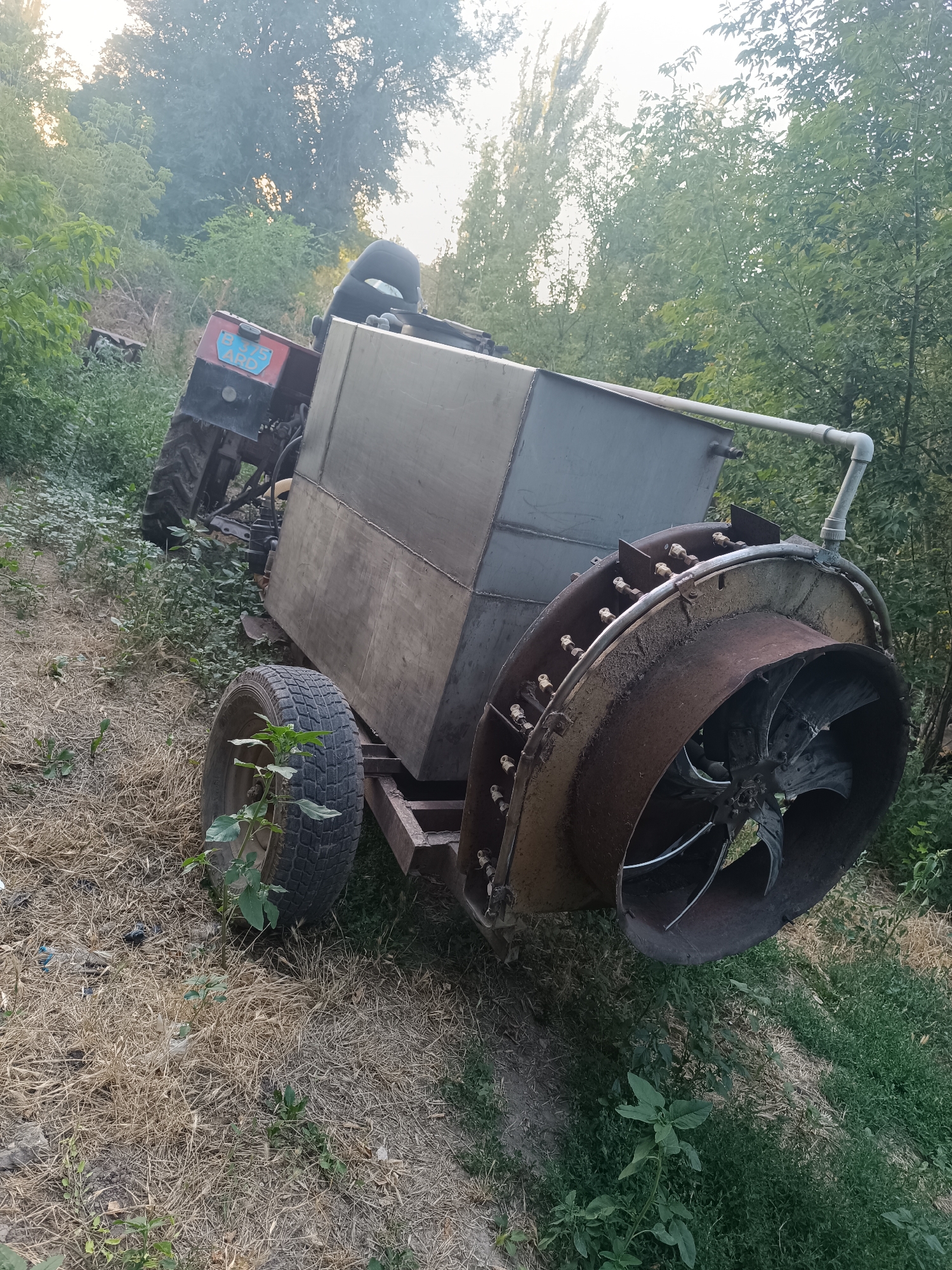 In the village of Baidibek Bi, Kazakhstan, an abandoned agricultural implement, likely a sprayer or atomizer, rests on overgrown dry grass and vegetation. The machine is connected to a small, weathered red tractor. The apparatus features a large, cylindrical metal tank mounted on a trailer chassis with a single, large rear wheel visible. A prominent feature is a substantial fan mechanism at the rear of the tank, encased in a rusty metal shroud. A white PVC pipe extends from the top of the tank. The scene is set amidst dense green foliage and trees under a bright, overcast sky, suggesting an outdoor, rural location that has been left unattended. There are no people present in the image.