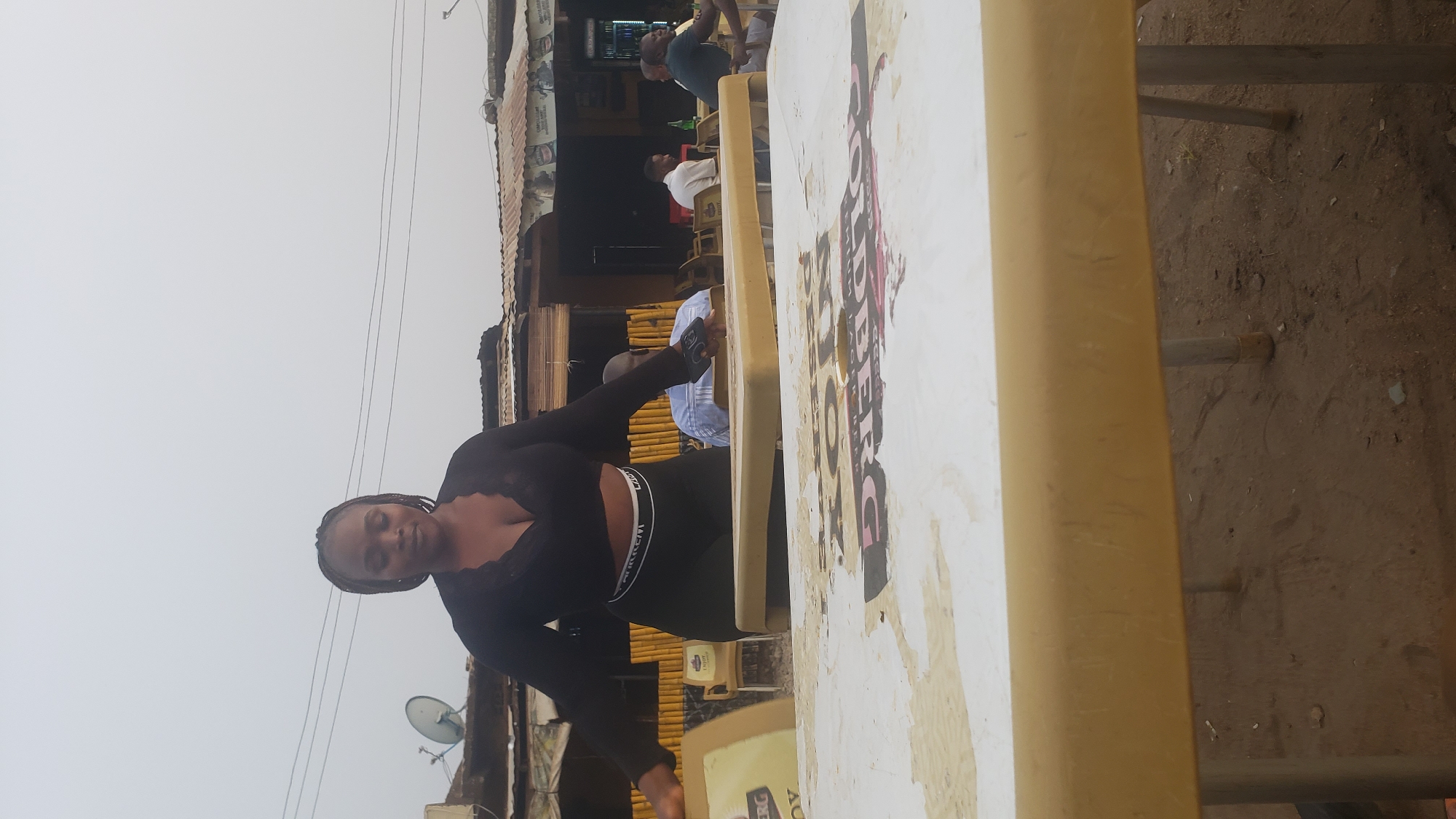 The image shows a woman wearing a black top and leggings standing outside a building. In the background, there are other people sitting at tables. There is a table in the foreground with an advertisement for Goldberg beer on it. Based on the presence of the woman, other people, and the beer advertisement, it appears to be a casual, outdoor setting in Narayi, Nigeria.