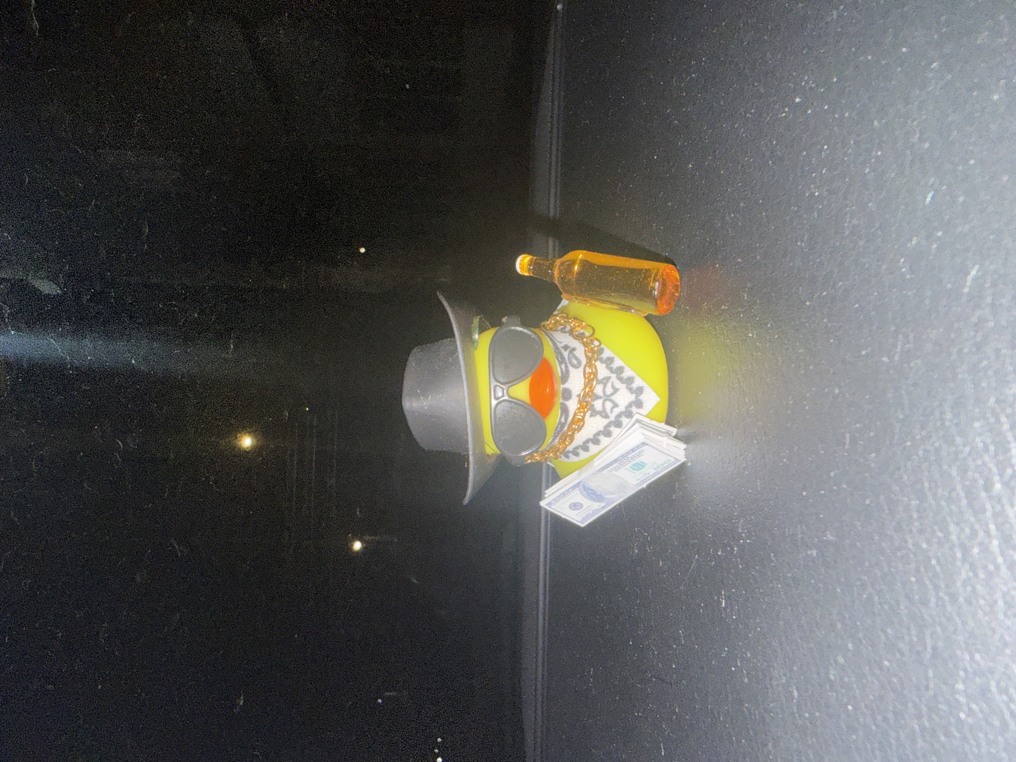A yellow rubber duck wearing sunglasses, a black hat, and a gold chain is seen in a photograph. It has a stack of cash resting on its chest and a miniature liquor bottle sitting on its back. The duck is positioned on a dark surface. The location in Elhovo, Bulgaria is not discernible from the image.