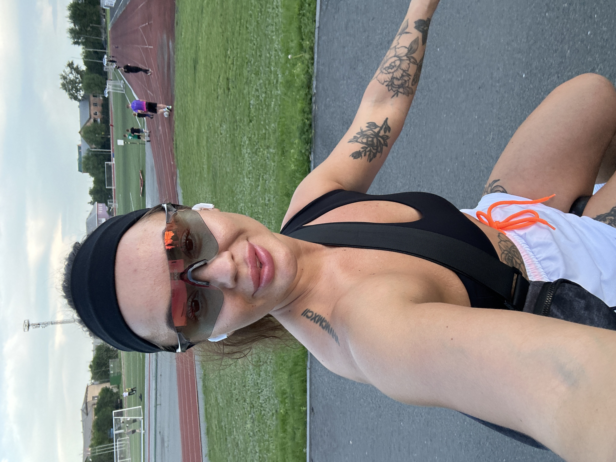 In Yuzhnouralsk, Russia, a woman with a black headband and sunglasses is posing for a selfie at an outdoor athletic track. She is wearing a black sports bra and white shorts with orange drawstrings. A black fanny pack is worn diagonally across her chest. She has several tattoos on her arms and shoulder blade, including Roman numerals on her left shoulder blade. In the background, a grassy field with a running track is visible, with several people engaged in various activities, including playing soccer and running. Buildings and a telecommunications tower are present in the distance under a cloudy sky.