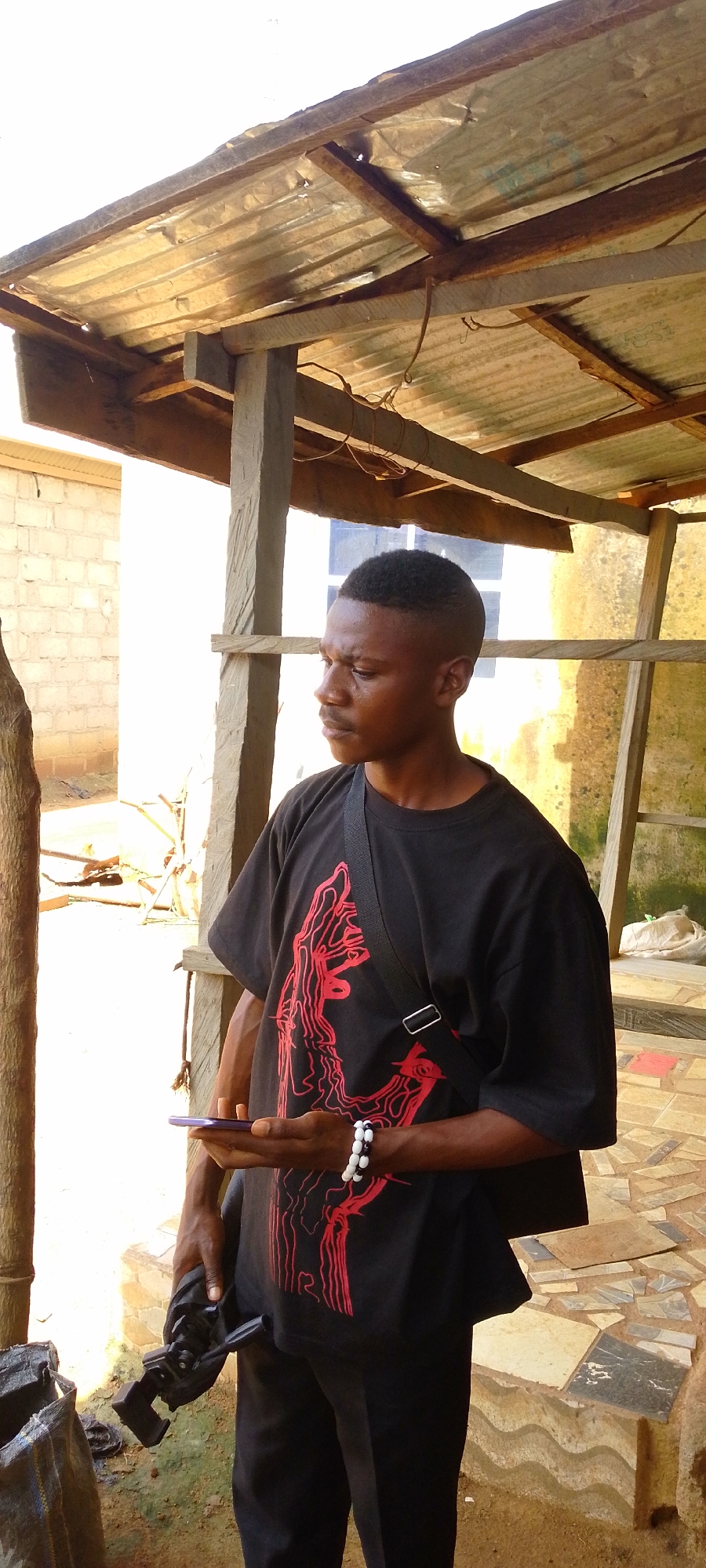 A young Black man is standing outdoors under a corrugated metal roof supported by wooden beams. He is wearing a black t-shirt with a red graphic design, black pants, and a black crossbody bag. He is holding a purple smartphone in his right hand and a black camera grip in his left hand. He is also wearing a bracelet made of black and white beads on his right wrist. The man is looking to his left, with a neutral expression.
The background shows a brick wall to the left and a yellow wall or building to the right. The ground is covered with rough-hewn stones and some dirt. Debris, including what looks like construction materials and a large plastic bag, is visible in the background. The lighting suggests it is daytime, possibly late afternoon, given the warm tones. The overall impression is of an outdoor, perhaps semi-constructed, area.