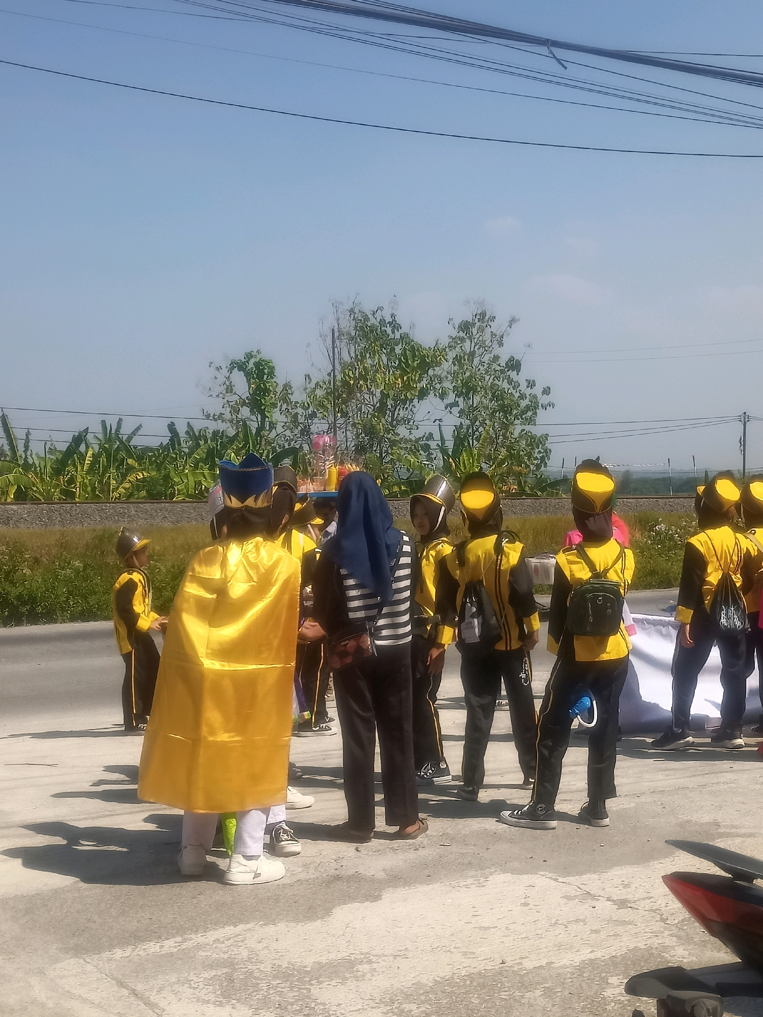 A group of children in yellow and black marching band uniforms with yellow hats stand in a line on a paved road under a bright, clear sky. One child in the front is wearing a large, shimmering golden cape over their uniform and a blue and gold crown-like hat. Behind them, several other children are visible, some wearing similar uniforms and hats, and one child is wearing a blue headscarf with a striped shirt. In the background, there is a line of banana trees, a stone wall, and a faint glimpse of a distant landscape. Power lines are strung across the top of the frame. The overall scene suggests an outdoor event or parade in Sumberlawang, Indonesia, likely on a sunny day.