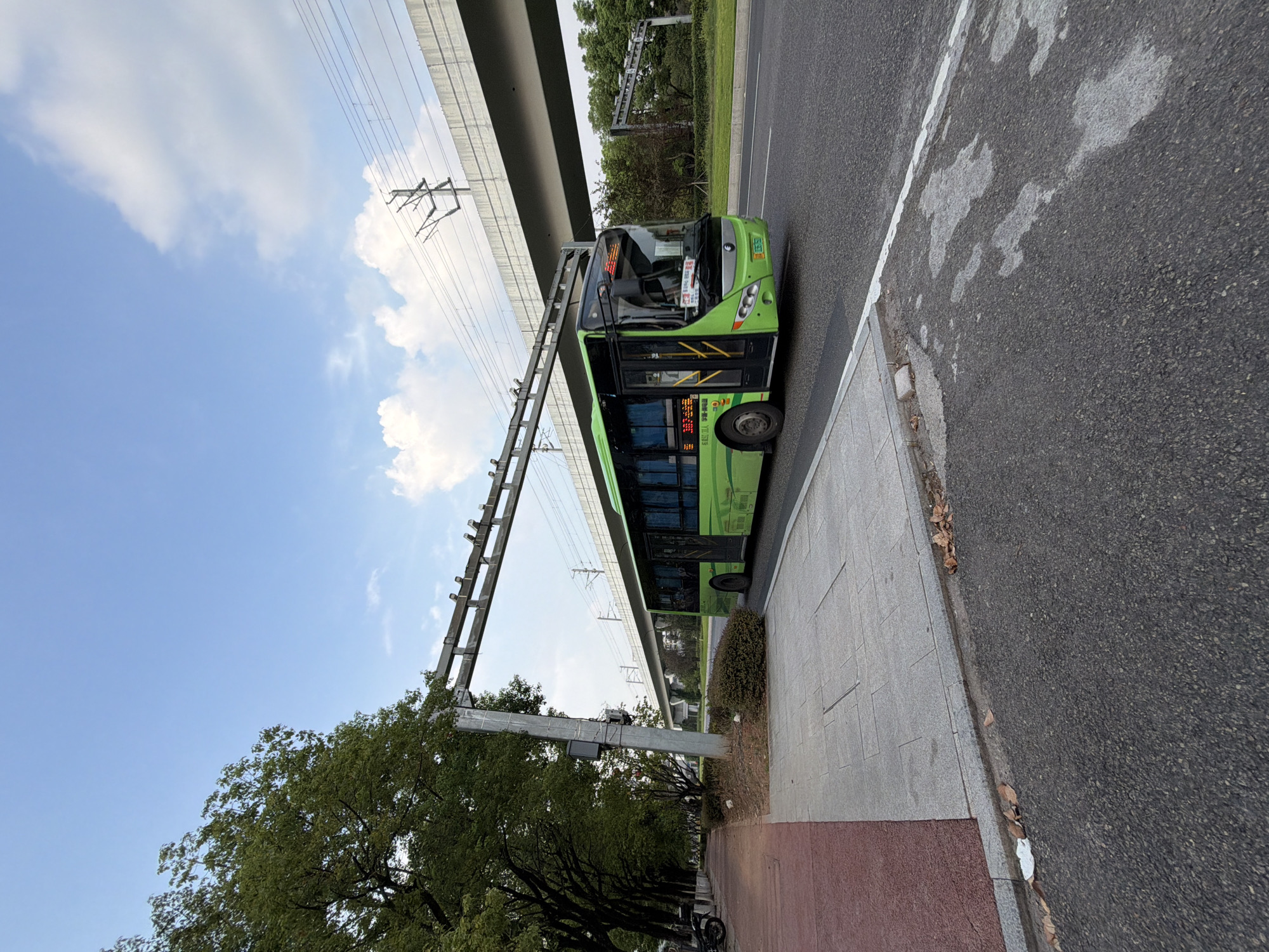 This image depicts a bright green bus driving on a road in Jiangjin, China. The bus is in motion and is positioned on the right side of the road. The foreground shows a paved sidewalk and a road with some debris, including fallen leaves. In the background, there is an elevated structure, likely a bridge or an overpass, with overhead power lines. Trees line the left side of the road, casting shadows. The sky is clear with some scattered clouds, suggesting it is daytime. There are no people clearly visible in the image. The scene appears to be an urban or suburban environment.
