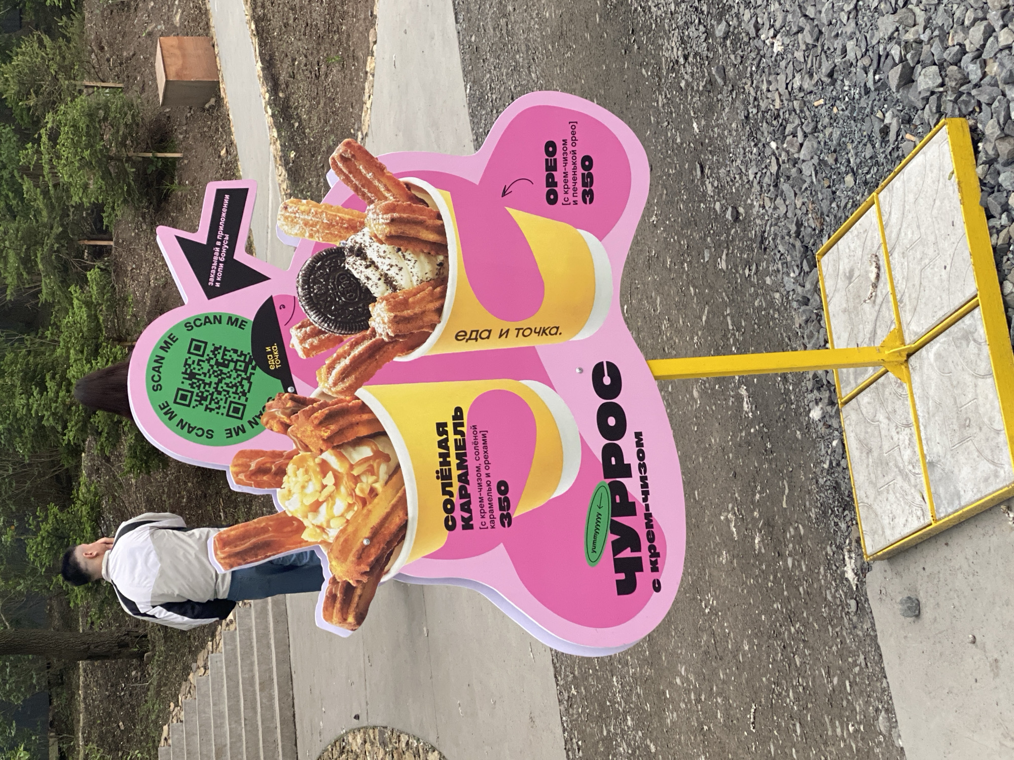 A close-up, eye-level, outdoor shot shows a pink and yellow sign advertising churros for sale. The sign has an abstract shape and features two illustrated cups of churros with toppings. The left cup has churros with caramel and nuts, labeled "Солёная КАРАМЕЛЬ" and priced at 350. The right cup has churros with cream cheese and an Oreo cookie, labeled "OREO" and also priced at 350. The main text on the sign, in large black letters, reads "ЧУРРОС с крем-чизом". A QR code is visible on the upper left of the sign, with the text "SCAN ME". To the right of the QR code, a black arrow points to text that reads "заказывай в приложении и копи бонусы".
In the background, a person wearing a white and black jacket and blue jeans stands facing away from the camera, looking towards a set of concrete stairs. The environment appears to be a park or outdoor recreational area with trees and gravel on the ground.