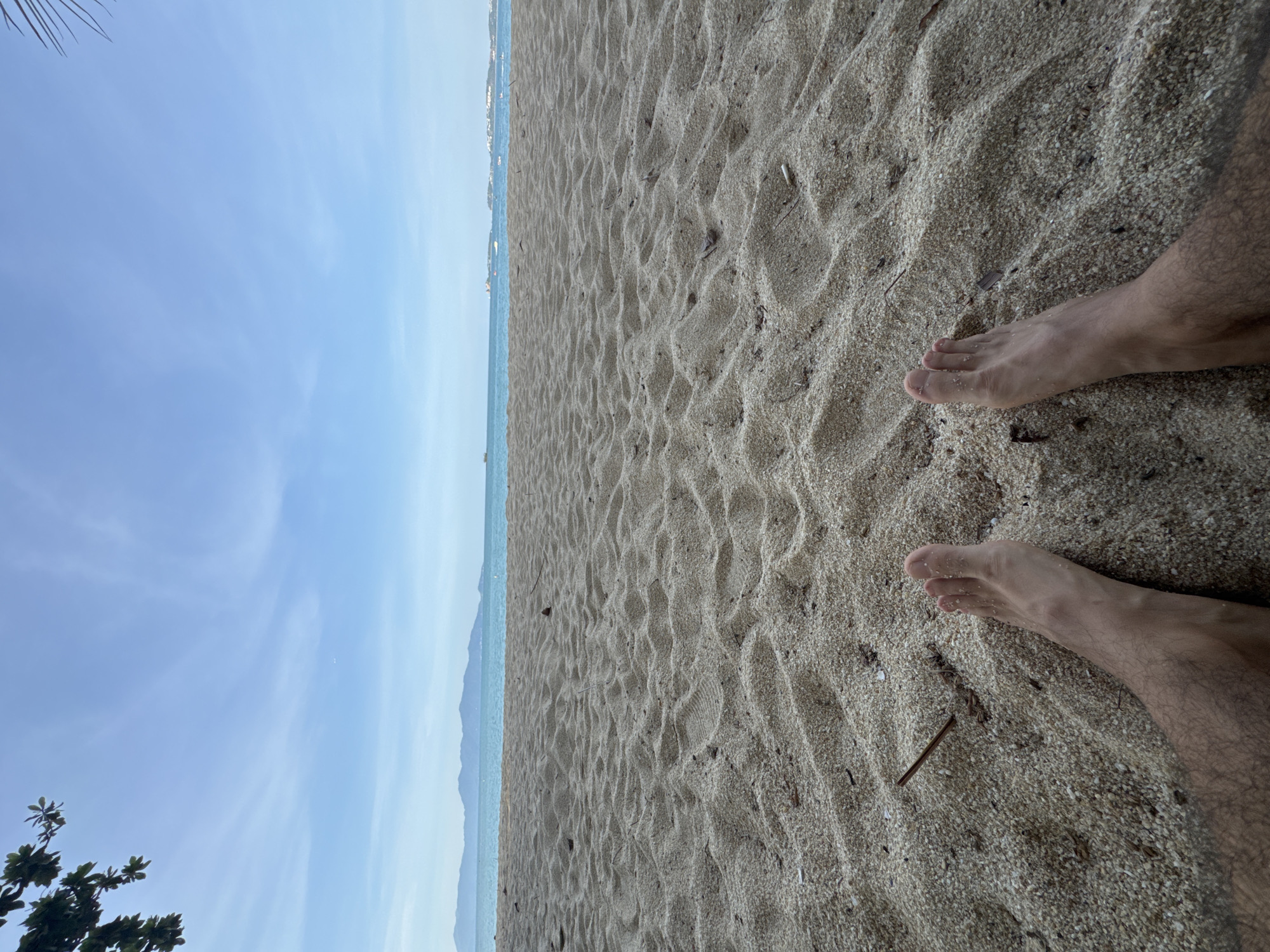 The image shows a person's bare feet and legs partially buried in the sand on a beach. The perspective is from the ground, looking out towards the ocean. The sand is light brown and textured with ripples. In the distance, the turquoise ocean meets a pale blue sky with some wispy clouds. A hazy mountain range is visible on the horizon. A few small twigs and debris are scattered in the sand. A portion of a leafy branch from a tree is visible in the upper left corner, and a palm frond is in the upper right corner. The scene is likely outdoors, on a beach in Ko Samui District, Thailand.