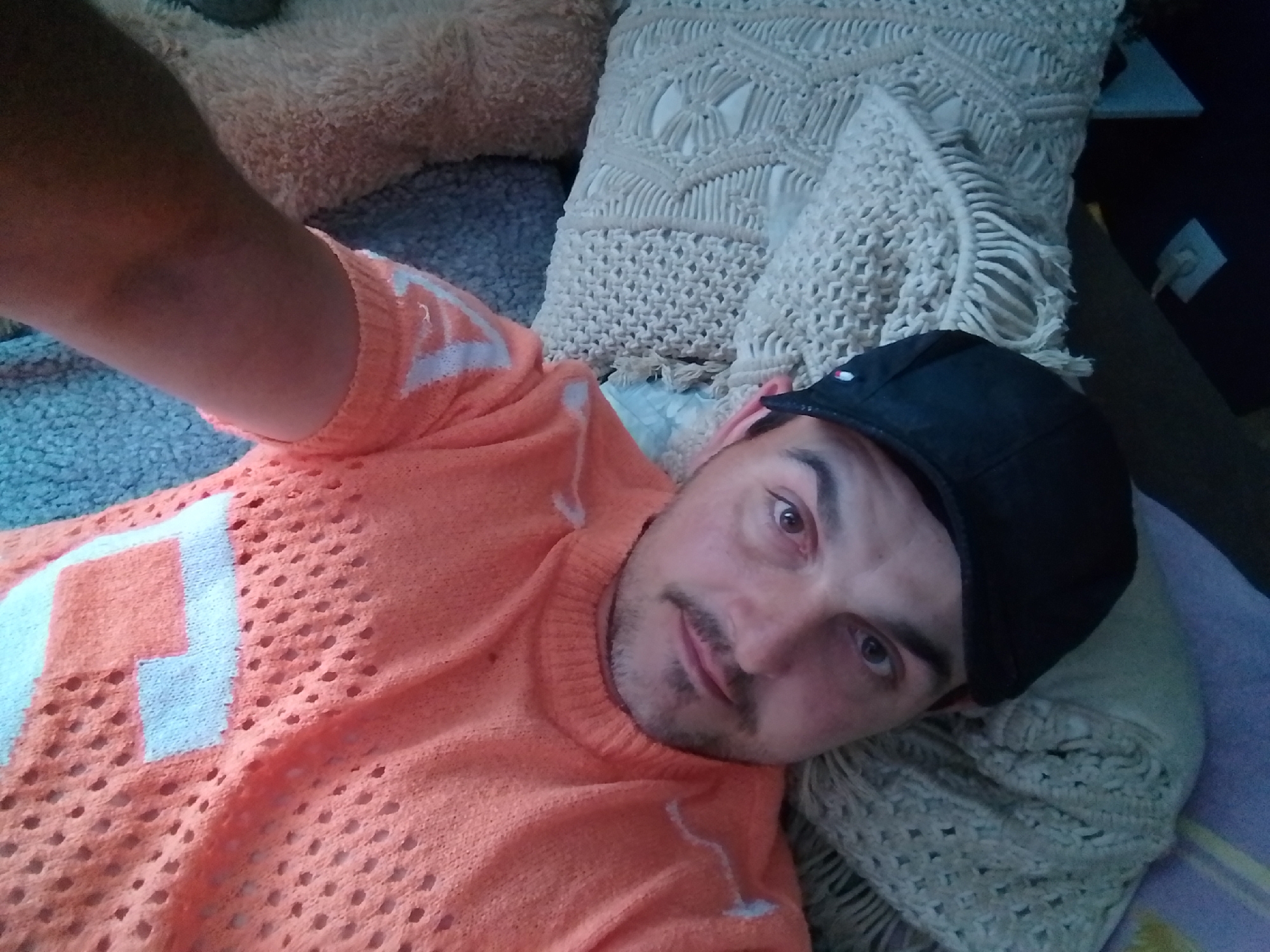 A man is lying on a bed, looking at the camera. He is wearing an orange sweater with white detailing and a black baseball cap. The man is of average build and has a mustache and stubble. The bed is covered with blankets and pillows, one of which has a macrame pattern. The room appears to be dimly lit. There is no indication of the man's location within the city of Wedel, Germany.