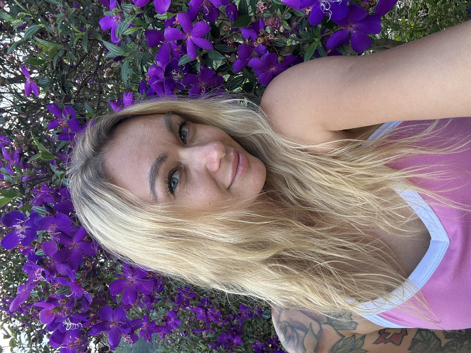 This is a close-up, selfie-style photograph of a woman lying down outdoors, with her head and upper body partially obscured by lush greenery and vibrant purple flowers. She has blonde hair and fair skin, and is wearing a pink tank top with white trim. Her eyes are blue-green, and she is looking directly at the camera with a slight smile. There are tattoos visible on her arm and shoulder. The purple flowers, likely Tibouchina, are in full bloom and form a natural backdrop. The lighting suggests it is daytime, and the overall atmosphere is relaxed and natural, hinting at pleasant weather. The image was taken in Thôn 3, Vietnam, adding a tropical context to the scene. The focus is on the woman and the surrounding flora, creating a serene and picturesque composition.