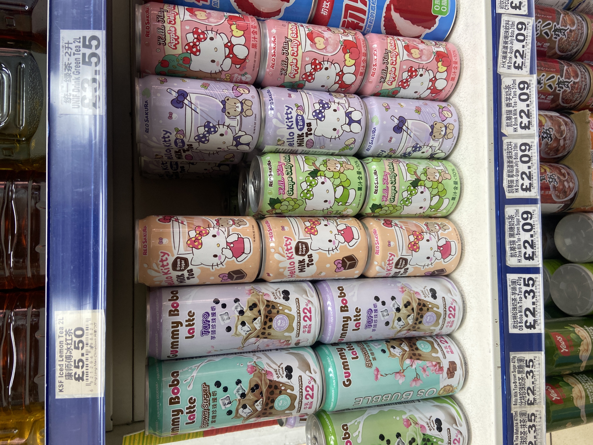 The image shows a shelf in a store, likely a Chinese supermarket in London's Chinatown, based on the product labels and price tags in GBP (£). The shelf is stocked with a variety of canned beverages. Dominating the scene are rows of "Hello Kitty" branded milk tea and jelly boba drinks in flavors such as apple, grape, taro, and brown sugar. There are also rows of "Gummy Boba Latte" drinks in taro and brown sugar flavors. To the left of the canned drinks, there are bottles of KSF Iced Lemon Tea and UNIF Drink Green Tea. To the right are other canned drinks, including HK brand fruit jelly drinks, as well as Yeo's brand drinks. Price tags are visible for various items, including Boba Milk Tea at £2.35, Hello Kitty drinks at £2.09, Iced Lemon Tea at £5.50, and Green Tea at £3.55. The shelf itself has a blue shelf edge.