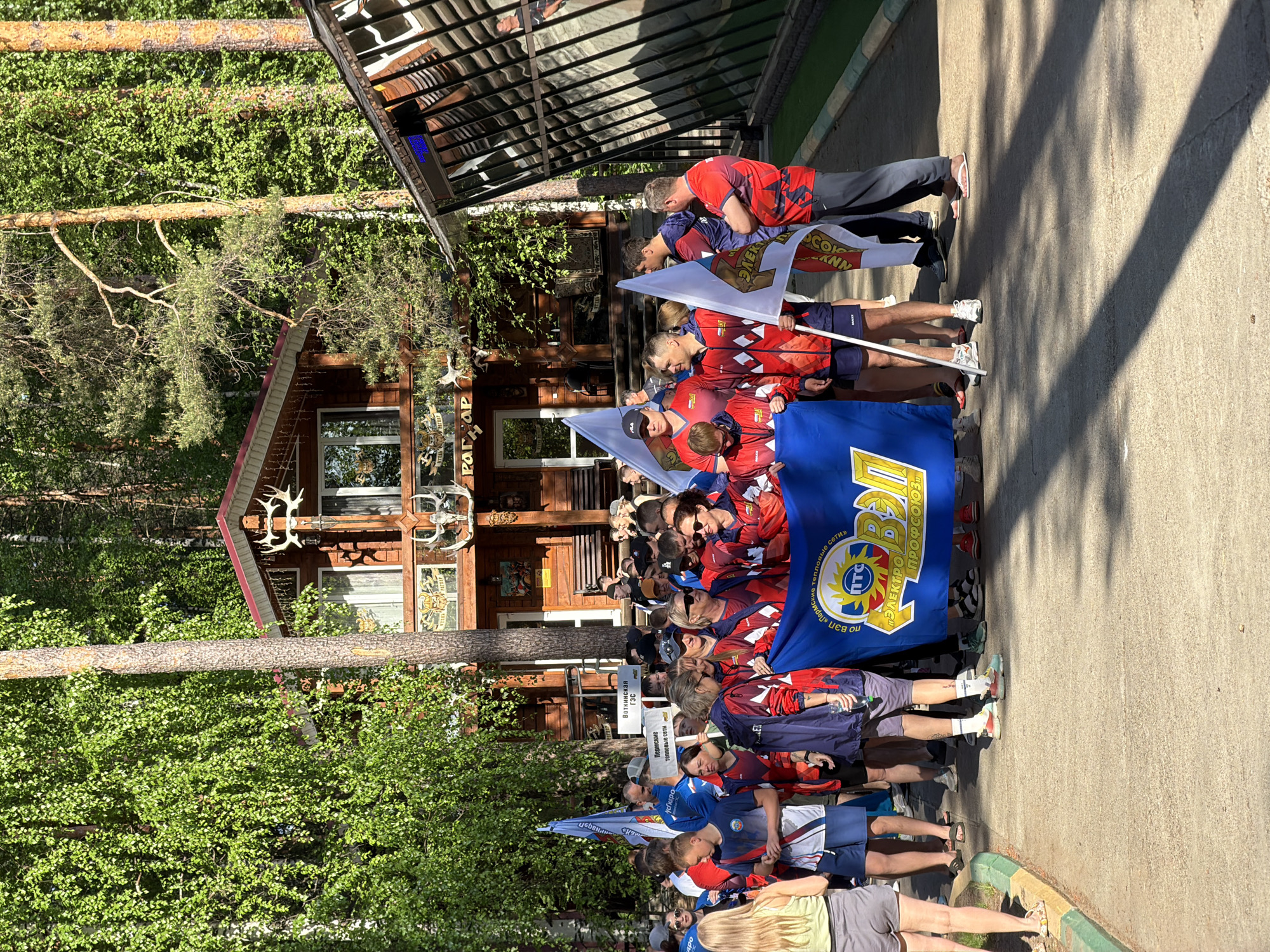 A large group of people stand along a concrete walkway in what appears to be Chaykovsky, Russia, judging by the Cyrillic text on flags and signs. They are carrying flags and banners, one of which is a large blue banner with the words "ELECTRO PROFSOYUZ" in Cyrillic. Some are wearing matching red and blue athletic wear. The group appears to be posing or participating in an event. Behind them is a wooden building decorated with antlers. Trees line both sides of the walkway. The overall environment suggests a park or recreational area.