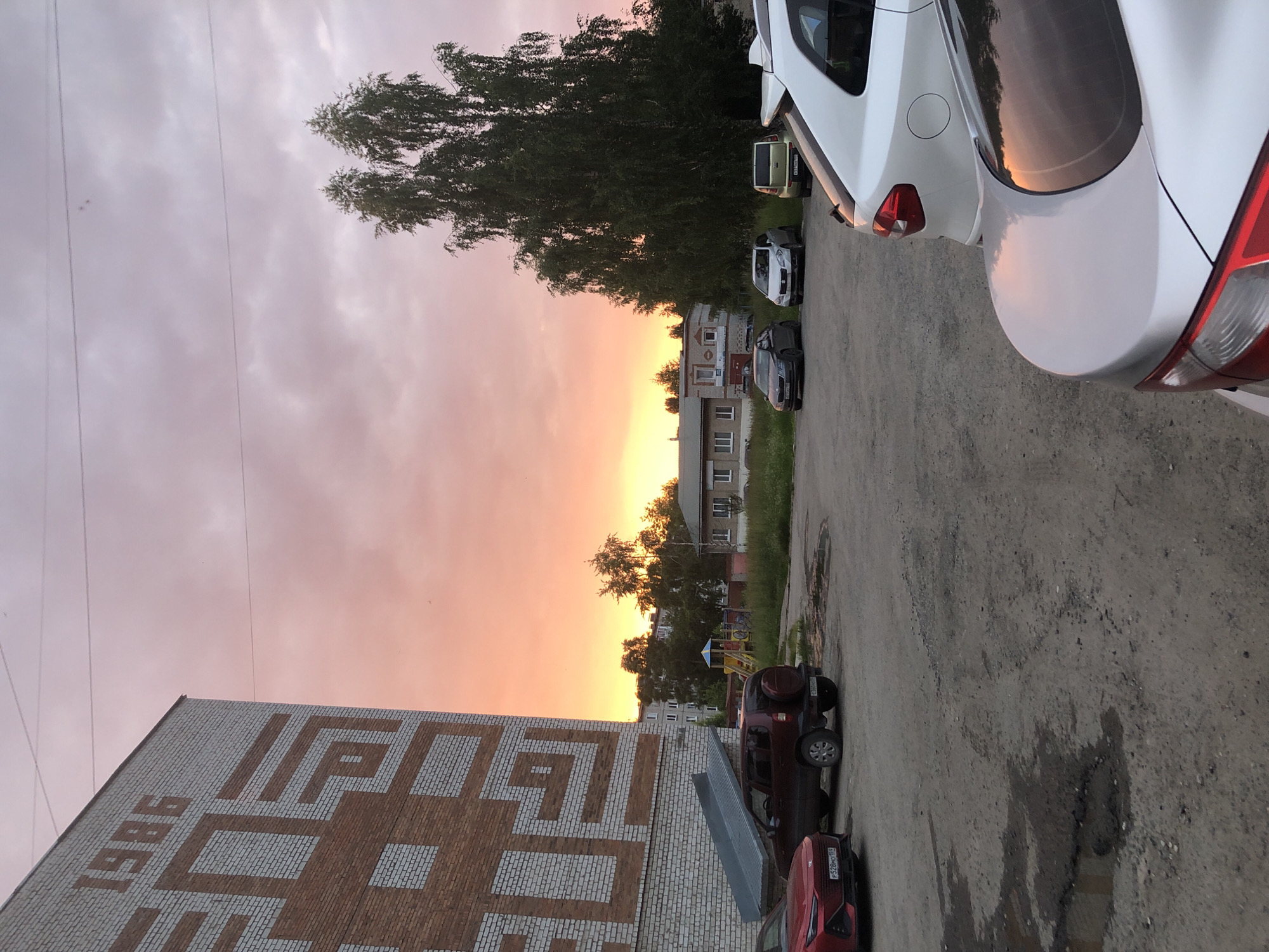 This vertical orientation media file depicts a sunset scene in a residential area, likely Solikamsk, Russia. The sky is filled with hues of pink and orange, indicating it is either dusk or dawn. The clouds are scattered, suggesting fair weather. In the foreground, a brick building is partially visible with large, stylized white and brown lettering that appears to spell "1986" and another word. Several cars are parked on an asphalt road beside the building, including a white car in the upper right, a dark SUV, and a red car at the bottom. Behind the cars, a line of trees and multi-story apartment buildings can be seen. A playground with yellow and blue structures is also visible amongst the trees. The overall atmosphere is peaceful and serene, capturing a quiet moment in a suburban or urban setting. There are no people visible in the frame.