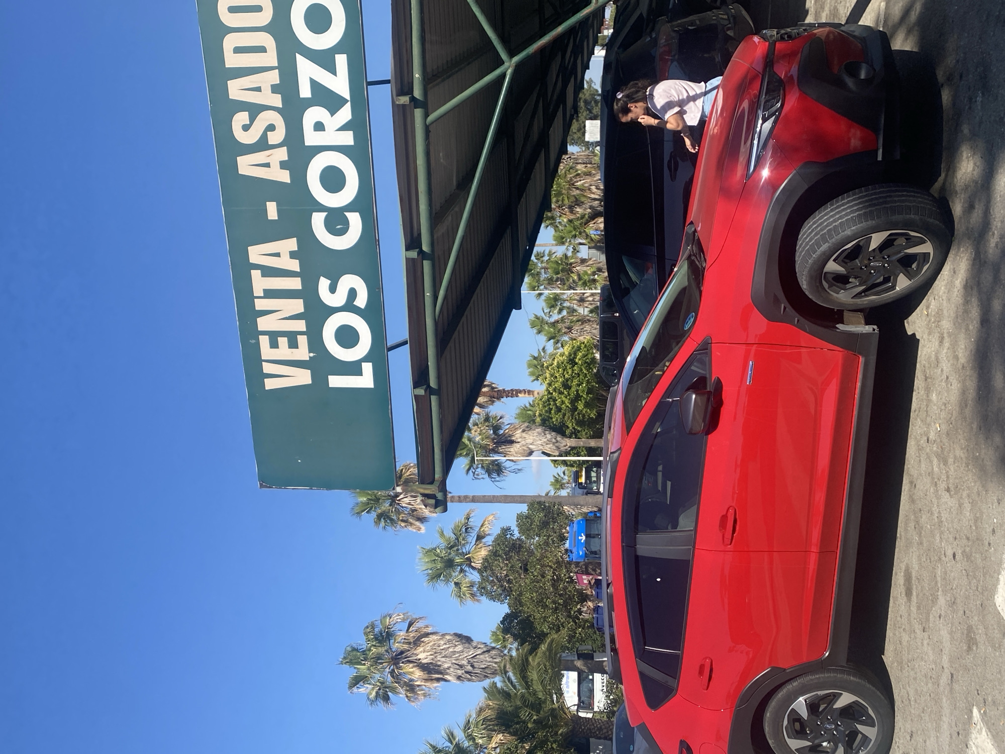 The image shows a red Subaru Crosstrek parked in an outdoor lot on a sunny day. A green sign with white text reads "VENTA - ASADO LOS CORZO". In the background, several palm trees are visible, along with other parked cars and a blue truck. A woman in a pink shirt and blue jeans is standing near a dark-colored SUV to the right of the red car. The scene appears to be in Pol. Ind. La Palmosa, Spain, as indicated by the sign, which suggests a commercial area or business.