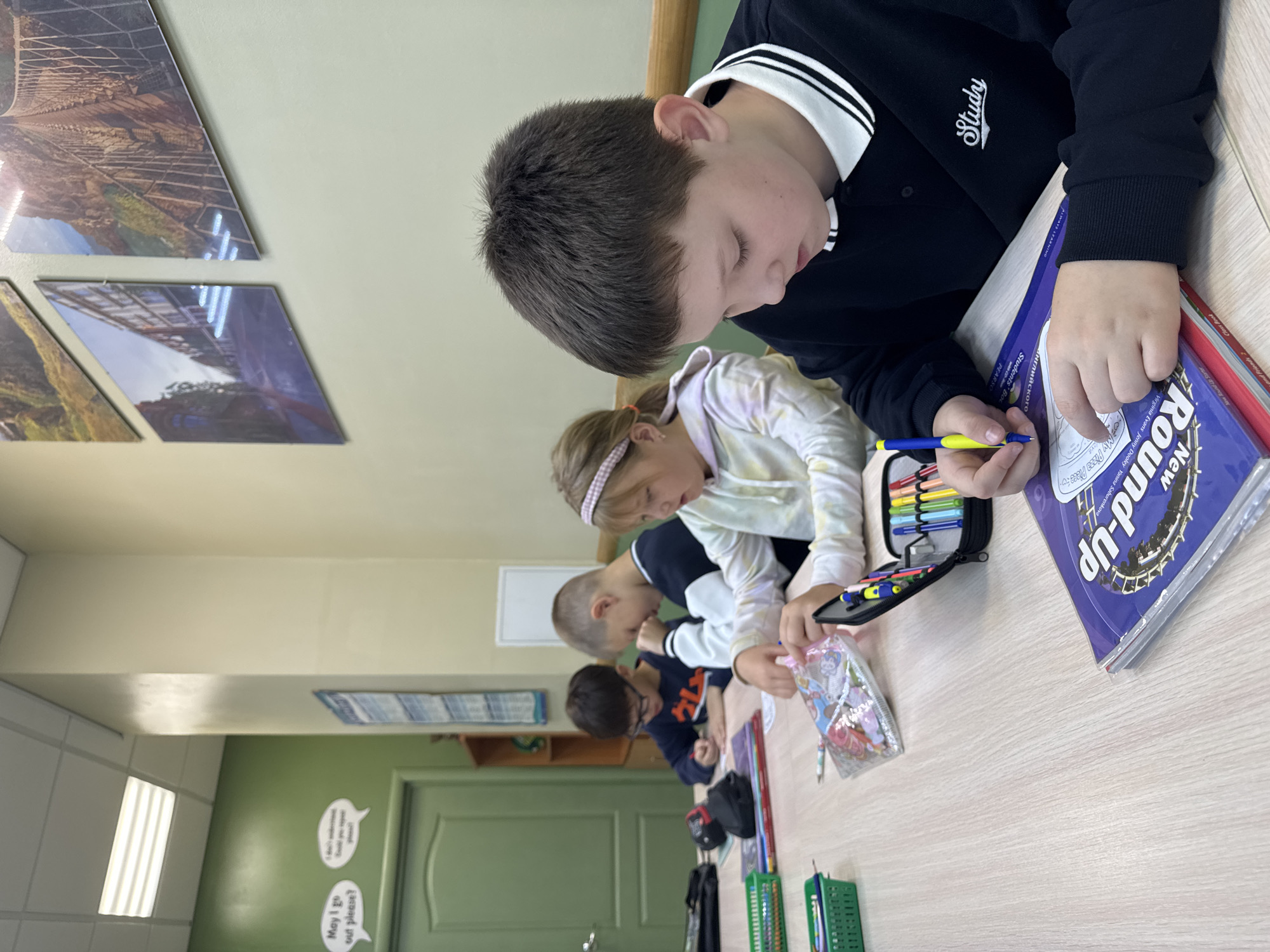 Four children are seated at a wooden table, engaged in activities that appear to be schoolwork or art. The child in the foreground on the right is a young boy with short, dark hair, wearing a black collared shirt with a white striped collar and the word "Study" embroidered on the left chest. He is focused on a book with a purple cover, holding a yellow and blue pen and writing on a piece of paper. To his left, a young girl with blonde hair, wearing a light-colored hoodie, is also writing or drawing. She has a pink headband and is looking down at her work. To her left, a boy with dark hair and glasses, wearing a dark blue sweatshirt, is leaning on his hand and looking at papers on the table. The boy furthest to the left, partially obscured, is also writing on papers.
The table is covered with various items including colorful markers in a pencil case, pens, notebooks, and art supplies in green plastic containers. The room has a light-colored ceiling with fluorescent lighting panels. The walls are painted in two tones: a pale yellow on the right and a vibrant green on the left. On the green wall, there are two cartoon-style speech bubbles; one reads "May I go out please?" and the other is partially obscured. Further down the green wall, there is a closed green door. On the pale yellow wall, there are several large framed pictures depicting scenic landscapes, including a suspension bridge and an urban street scene. A wooden shelf unit with books and other items is visible in the background on the left. The overall impression is that of a classroom or a study area.
The scene is not identifiable as being specifically located in the city of Perm, Russia. There are no discernible landmarks, signs, or architectural features that would indicate a specific city or country.