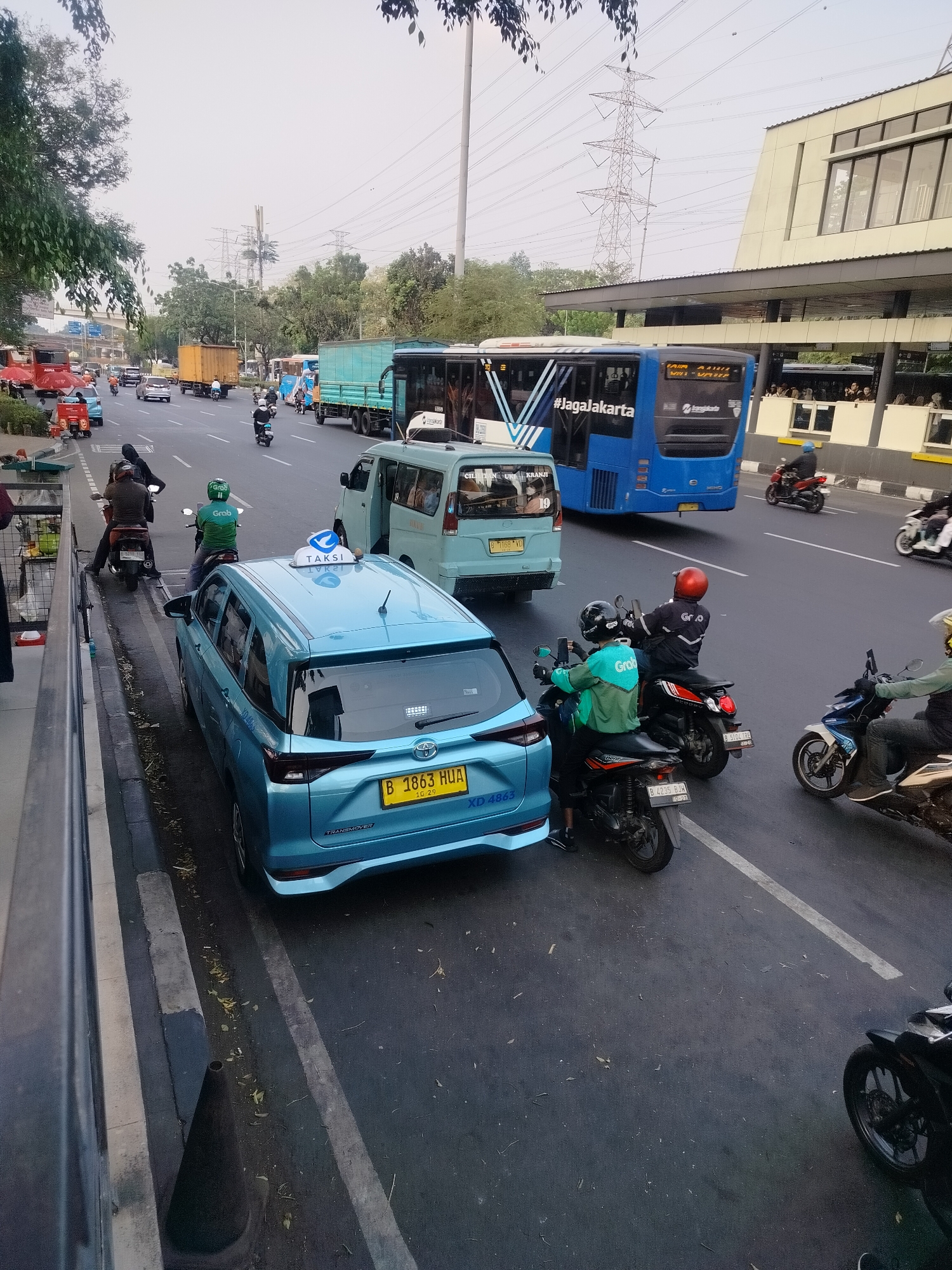 The image depicts a busy street scene in East Jakarta, Indonesia, likely during the day. In the foreground, a light blue taxi with a "TAKSI" sign on its roof is stopped, its rear facing the camera. Behind the taxi, several motorcycles are visible, some carrying passengers. One motorcycle has a rider wearing a green jacket with "Grab" logo, suggesting a ride-sharing service. Further back, a light blue passenger van and a large blue bus are on the road. The bus has "#JagaJakarta" and "2JagoLakarta" written on its side, indicating it's part of Jakarta's public transportation system. The background shows power lines, trees, and a modern building with glass windows. The road is paved and marked with white lines. The overall impression is one of urban traffic congestion.