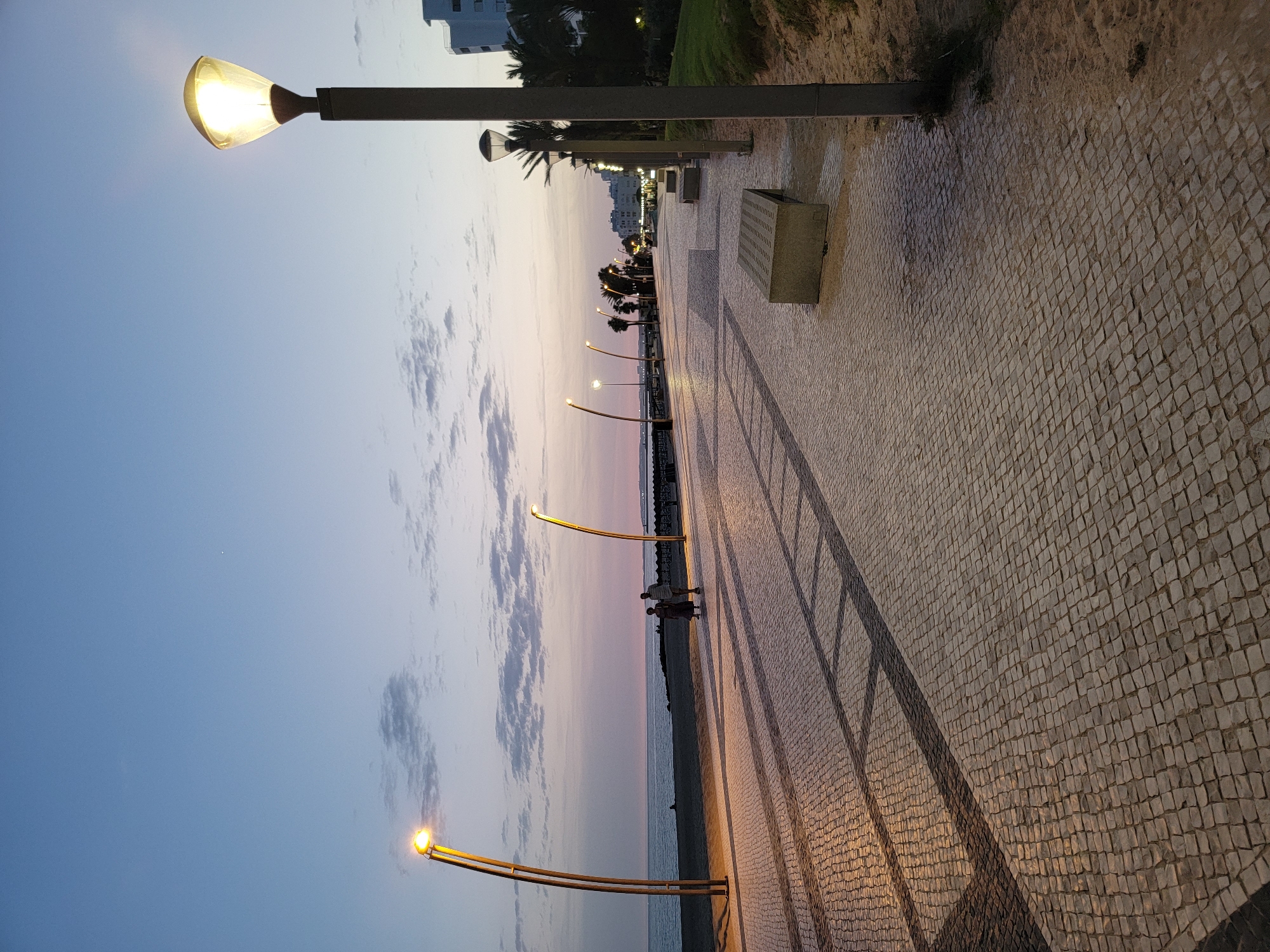 The image shows a wide, paved promenade at dusk in Quarteira, Portugal. The promenade is lined with modern, curved lampposts, some of which are illuminated, casting long shadows across the cobblestone walkway. In the distance, the ocean is visible, with a gentle shoreline and a few indistinct figures walking along the beach. A couple is strolling on the promenade. Several concrete benches are placed along the path. Beyond the promenade, there are palm trees and buildings, suggesting an urban coastal setting. The sky is a gradient of pale blue and pink, indicating the transition from sunset to twilight, with a few scattered clouds. The lighting from the lampposts suggests artificial illumination is beginning to dominate as natural light fades.