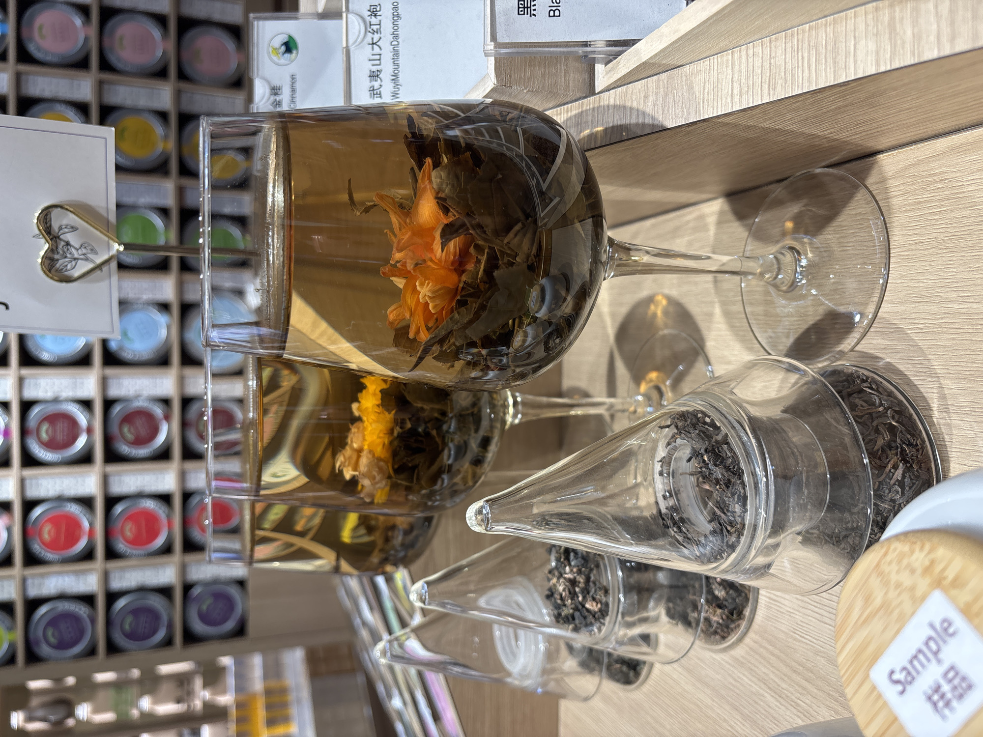 The image depicts a retail display of tea in what appears to be a store in Guangzhou City, China. Two clear, stemmed glasses filled with blooming tea (tea leaves wrapped around a dried flower) are prominent. The tea has been steeped, creating a light amber liquid. Behind these are several cone-shaped glass containers filled with loose tea leaves. A label with the word "Sample" is visible near the bottom right corner. In the background to the left, are numerous small containers of tea displayed in a grid pattern. Some of the tea canisters have labels in both Chinese and English. The setting appears to be a brightly lit retail space, possibly a tea shop or a specialty foods store.