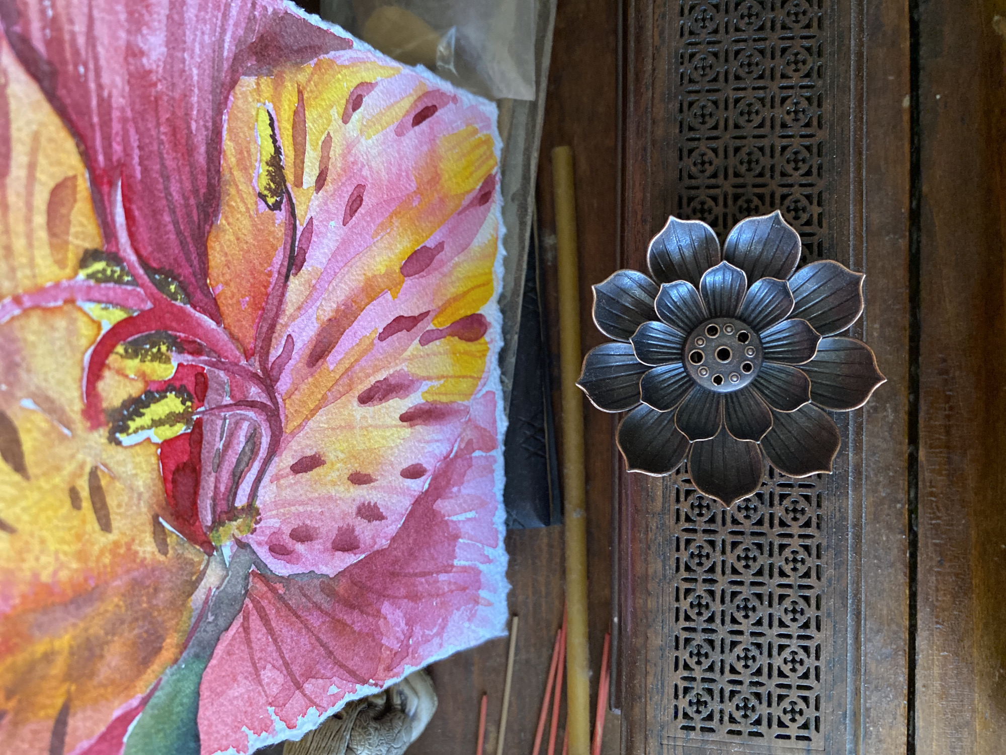 The image presents a still life arrangement on a dark wooden surface. To the left, there's a watercolor painting of a flower, predominantly in shades of red, yellow, and pink. The painting appears unfinished, with visible brushstrokes and a slightly torn edge. To the right of the painting, there are objects including a bronze-colored flower-shaped incense holder with a patterned center and multiple petals sitting on a wooden surface.
There is no indication that this image was taken in Izhevsk, Russia.