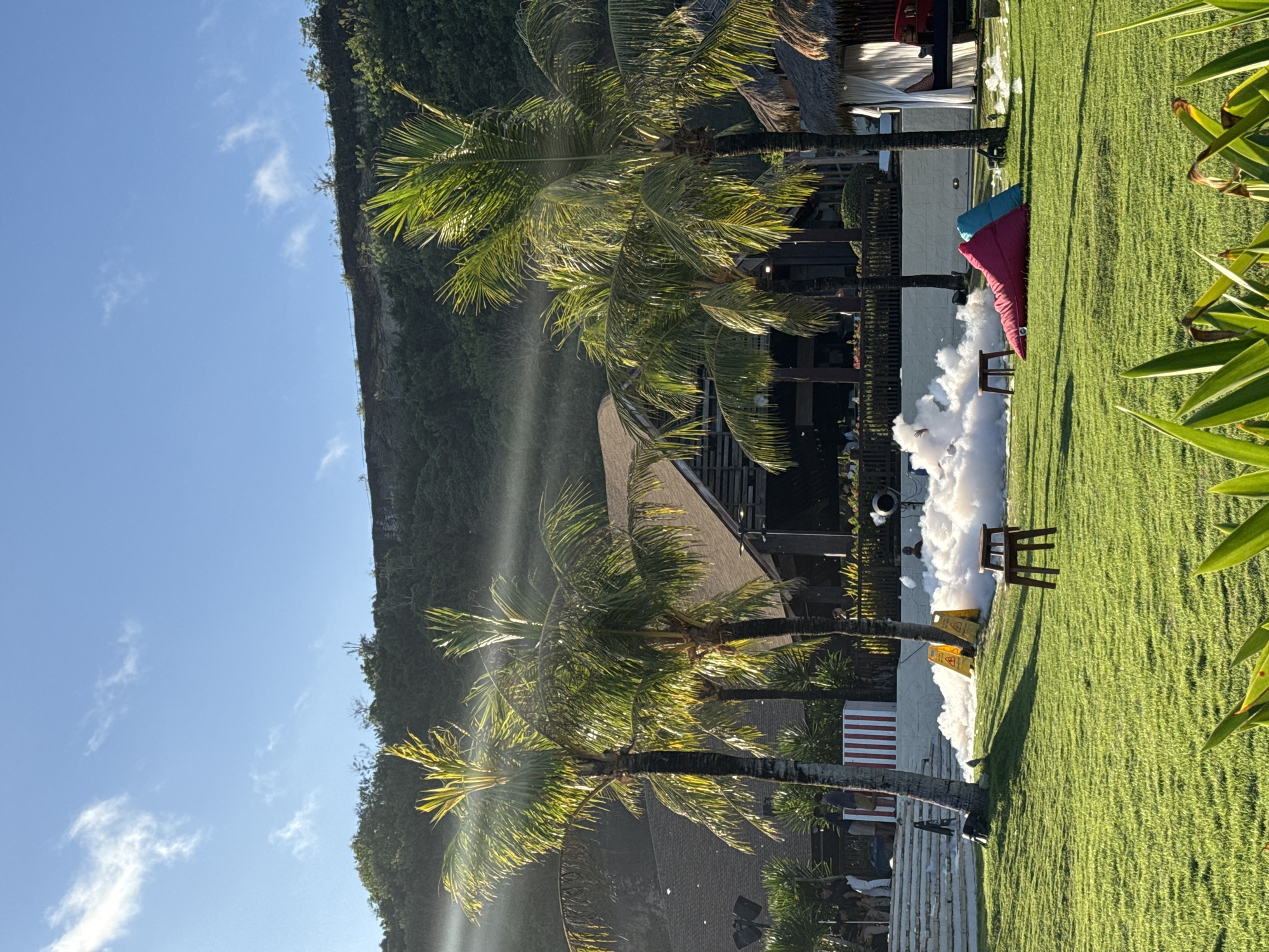 The image shows an outdoor event taking place in Kuta Selatan, Indonesia. Palm trees are prominent in the foreground and background with green vegetation. There is a foam machine active and dispensing white foam onto the green lawn. Two yellow caution signs are placed near the foam. A small wooden stool sits on the grass, and there are red and blue pillows placed against a wall. A cliff covered in vegetation is visible in the left of the image. The sky is clear with some clouds. Buildings with thatched roofs and an outdoor stage with lighting equipment can be seen among the palm trees. People are blurred in the background.
