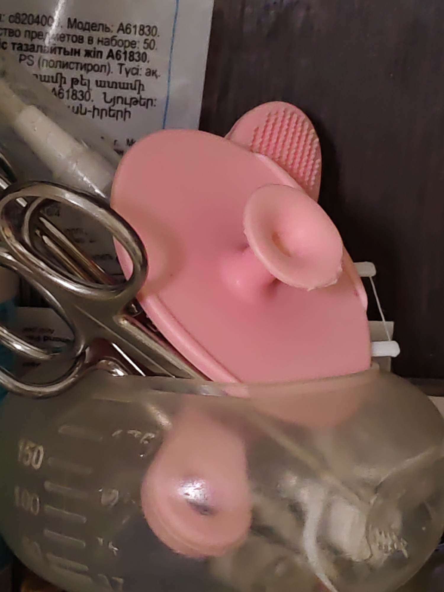 The image shows a close-up of various objects, including scissors, a pink silicone facial cleansing brush, and a clear plastic bottle with measurement markings. The background is dark and out of focus. Text is visible on a label attached to one of the items, with some of the text in Russian and Armenian. The text indicates a model number "A61830" and mentions "50" items in a set. There is no discernible indication of the location being Rostov-on-Don, Russia, nor is there any mention of people, actions, or interactions. The scene appears to be an indoor setting, likely a tabletop or shelf.