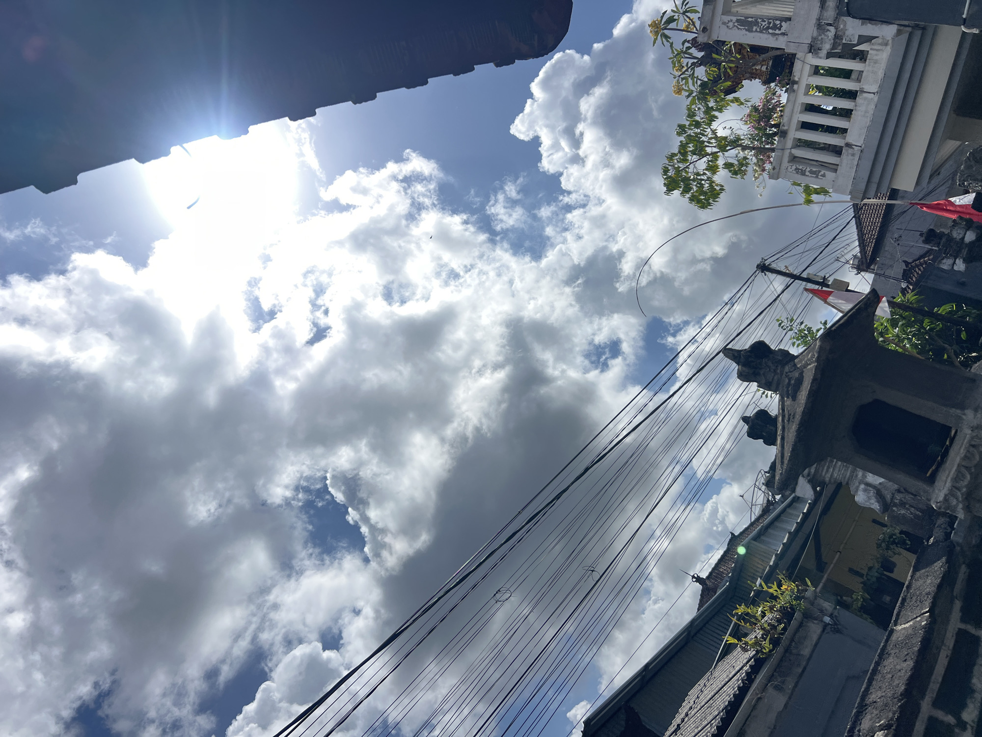 The image depicts a view looking upwards at a cloudy sky, with the sun intensely bright and positioned in the upper right quadrant, causing a lens flare. The foreground features a street scene in Denpasar, Indonesia, characterized by overhead power lines, a dark, textured building or roofline on the right, and rooftops of buildings on the left. Partially visible structures include what appears to be a small, decorative shrine or monument in the lower center, with intricate stonework. A white balcony with a railing is visible on the right, adorned with plants. Two Indonesian flags, one larger and one smaller, are partially visible, attached to poles. The overall impression is an outdoor urban environment under a dynamic sky.