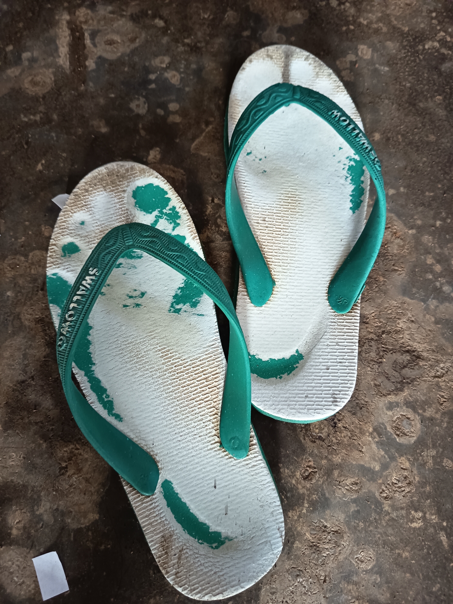 A pair of worn, white and green flip-flops with the brand name "SWALLOW" visible on the strap are lying on a textured, dark brown, dirt or concrete surface. The flip-flops show signs of wear with green paint chipped off in several areas, revealing the white material underneath. The left flip-flop is positioned at an angle, with the toe pointing towards the bottom left of the frame, and the right flip-flop is positioned more centrally, with the toe pointing towards the top right. The background surface has an uneven texture with lighter patches and darker, circular indentations, suggesting it is an outdoor ground surface in Krampilan, Indonesia.