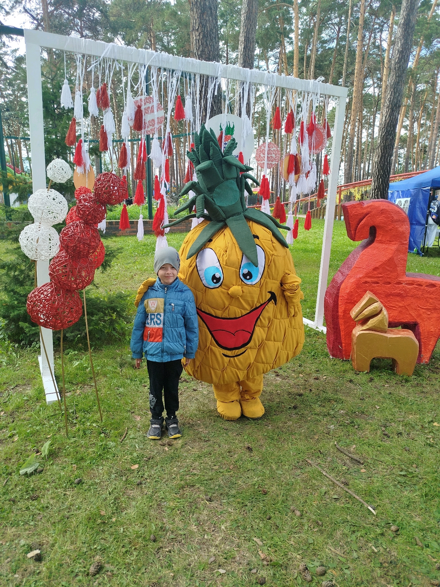 In Zavodoukovsk, Russia, on a grassy outdoor area with scattered pinecones, a young boy stands next to a person in a pineapple mascot costume. The boy, wearing a blue jacket, gray hat, and black pants, is positioned to the left of the mascot. The pineapple costume is large and yellow, with a green leafy headpiece, large blue eyes, and a wide red smile. Behind them, a white frame decorated with numerous red and white tassels hangs, and to the right, there are two carved wooden horse figures, one large and red, the other smaller and gold. In the background, tall pine trees are visible, and a blue tent with people around it is situated further back. The overall setting appears to be a public event or festival.