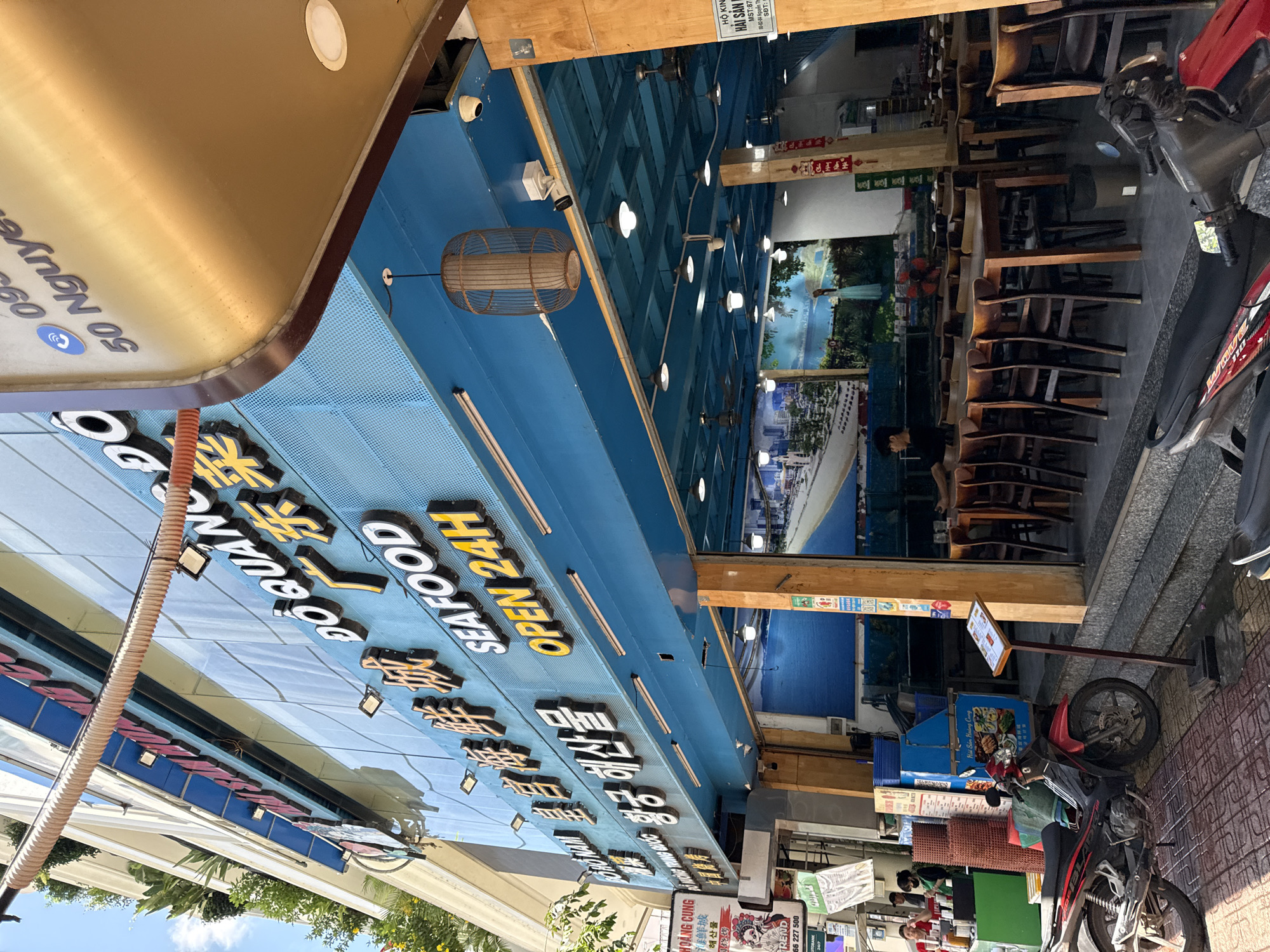 The image shows a street view of a seafood restaurant in Nha Trang, Vietnam. The restaurant's facade features prominent signage with Chinese and Vietnamese text, including "DO QUANG DO 广东 SEAFOOD OPEN 24H." A large gold-colored canopy is visible above the entrance. The restaurant appears to have an outdoor seating area with wooden tables and chairs, illuminated by ceiling lights and a decorative birdcage-style lamp. Inside, a mural depicting a coastal landscape is visible. A red and black motorcycle is parked on the sidewalk in front of the restaurant, near a menu stand. Several individuals are present, including a person in a black shirt inside the restaurant and two people standing near a green counter outside. The street is paved with patterned tiles, and trees with green foliage are visible in the background, suggesting a sunny day. The overall impression is that of a bustling urban environment focused on food service.
