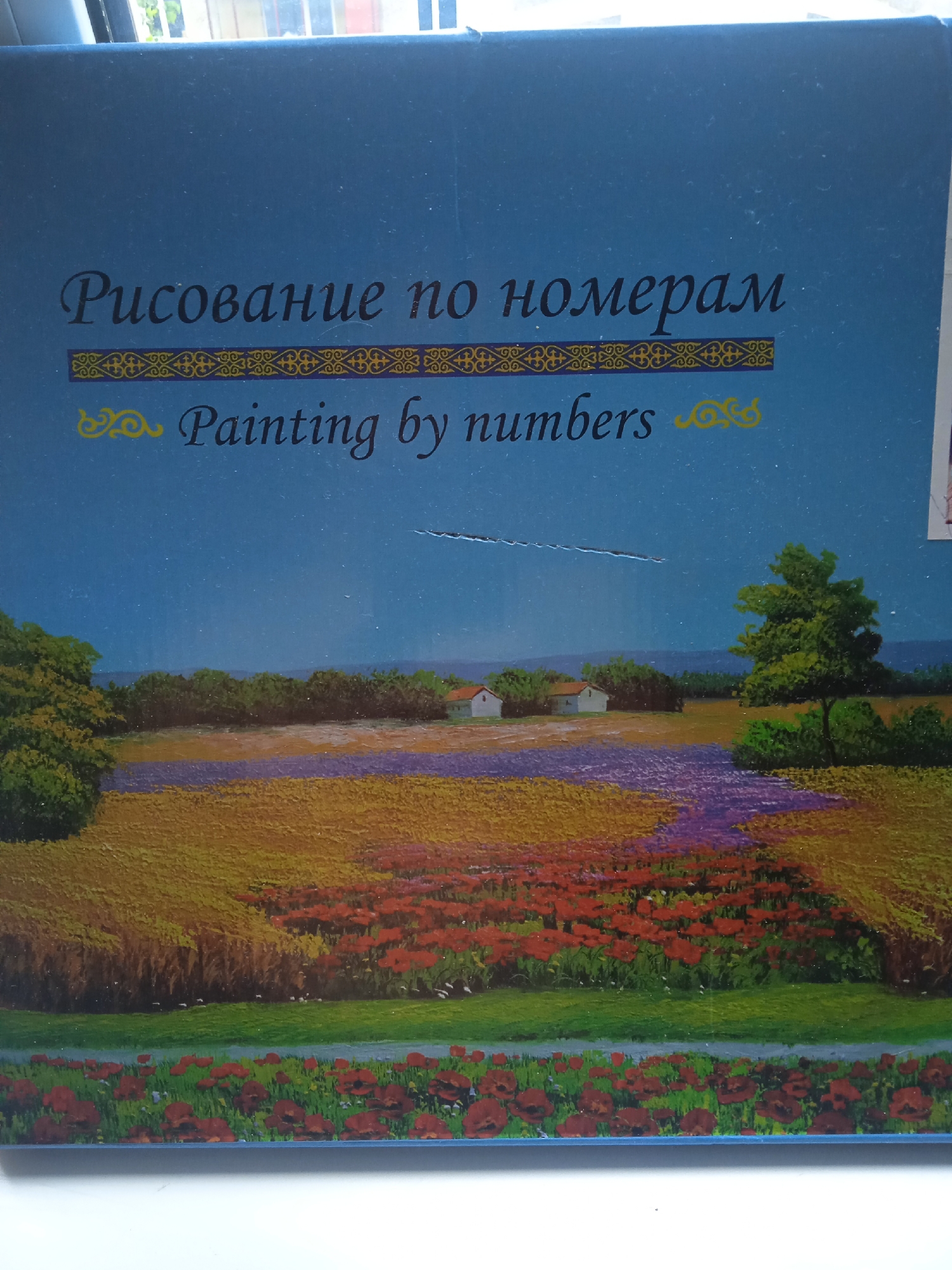 The image is a close-up of a box for a "Painting by numbers" kit. The box has a blue background with text in Russian and English. The Russian text reads "Рисование по номерам" and the English text reads "Painting by numbers". Below the text is a picturesque landscape scene, likely depicting the image to be painted. The scene features rolling hills with fields of yellow and purple flowers, a dirt path, a small white house with a red roof, and trees. The bottom edge of the box has a blue border. There is no visible indication of the location being in the city of Krasnoyarsk, Russia, or any specific geographical location. The image focuses on the product packaging and its illustrative content, not a real-world scene within a city.