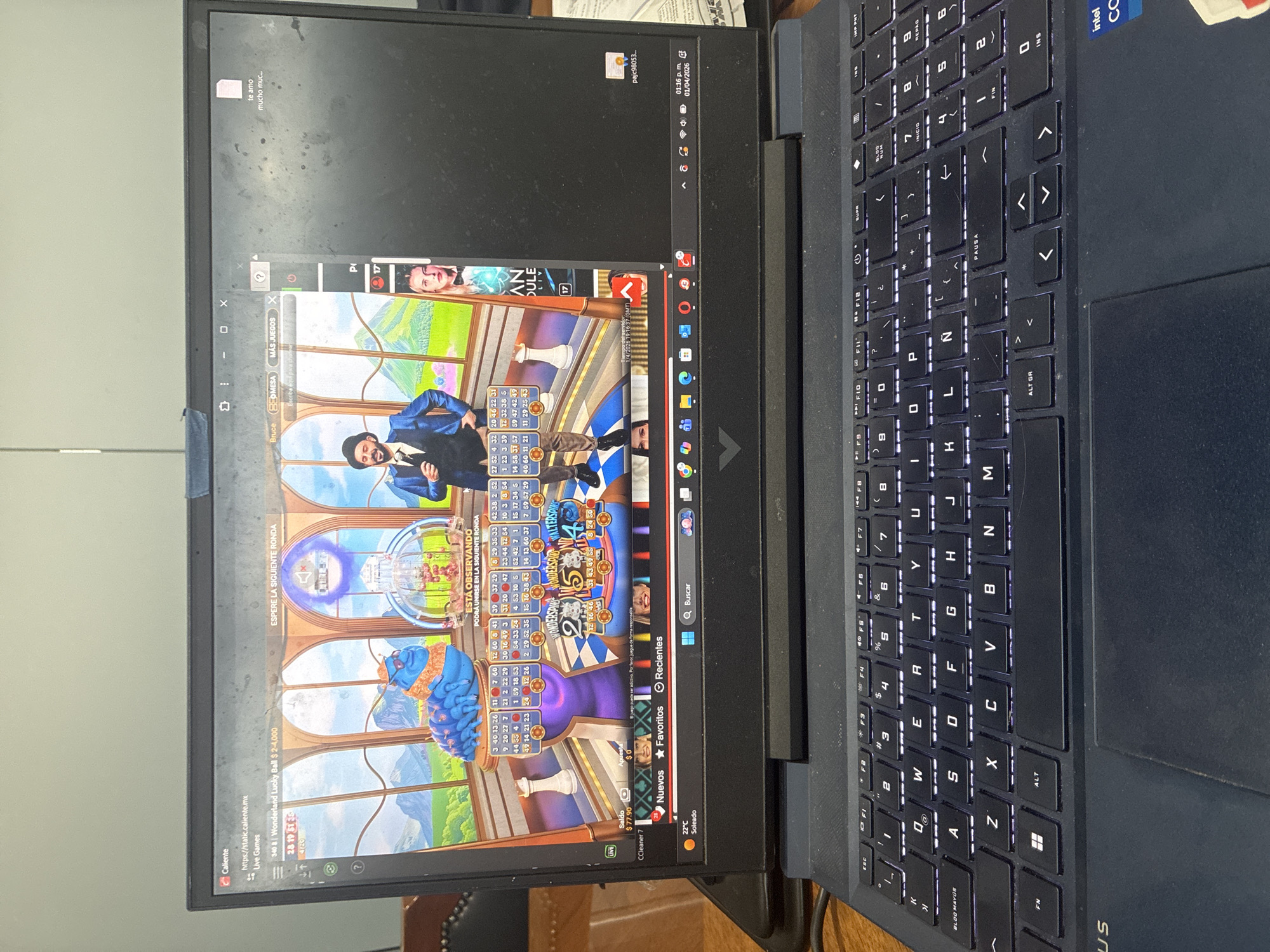 The image depicts a laptop screen displaying an online casino game, "Wonderland Lucky Ball." The game interface shows a spinning wheel, number displays, and cartoonish characters. Text on the screen indicates "Espere la siguiente ronda" (Wait for the next round). Various browser tabs and application icons are visible along the edges of the screen, including "Caliente" (a gambling website), "CCleaner 7", and a file named "pajc98053." The operating system's taskbar is also visible with a date, time and weather. The laptop itself has a black keyboard with illuminated keys and an Intel Core sticker on the top right corner. Based on the language displayed ("te amo mucho muc...") and the "Caliente" tab, it is most likely being used in Mexico. Without further evidence or information about the location or people involved, a precise description for Cuautitlán Izcalli, Mexico cannot be given.