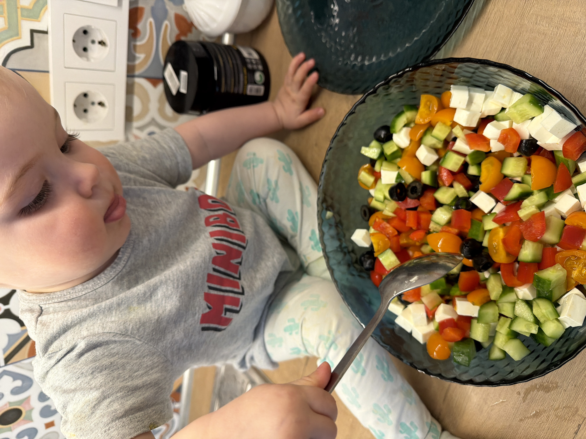 A baby wearing a grey t-shirt and patterned pants sits on the floor next to a bowl of salad. The salad contains cucumbers, tomatoes, olives, and feta cheese. The baby is holding a spoon in one hand and reaching towards the salad with the other. A black container is beside the salad bowl. A power outlet is visible in the background, as well as patterned floor tiles. The setting appears to be indoors. The exact location in Votkinsk, Russia, cannot be determined based on the image.