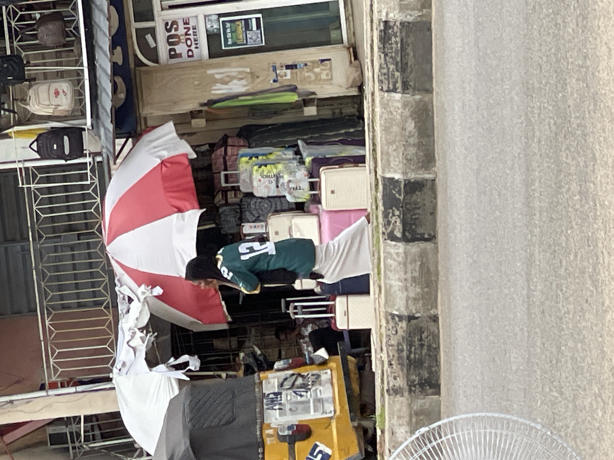 In a city scene, a woman wearing a green jersey with the number 12 and a headscarf walks past a stall filled with luggage. She is carrying a black scarf. The stall displays a variety of suitcases in different colors and sizes, some stacked high. A red and white umbrella is partially visible over the stall. To the left of the stall, a yellow auto-rickshaw is parked, its rear visible with license plates. In the background, a building has a balcony with a railing and several backpacks hanging from it. A sign on the building reads "POS DONE HERE". The scene appears to be on a street with a concrete barrier separating the road from the stall area. A white oscillating fan is visible in the foreground on the left side of the image.